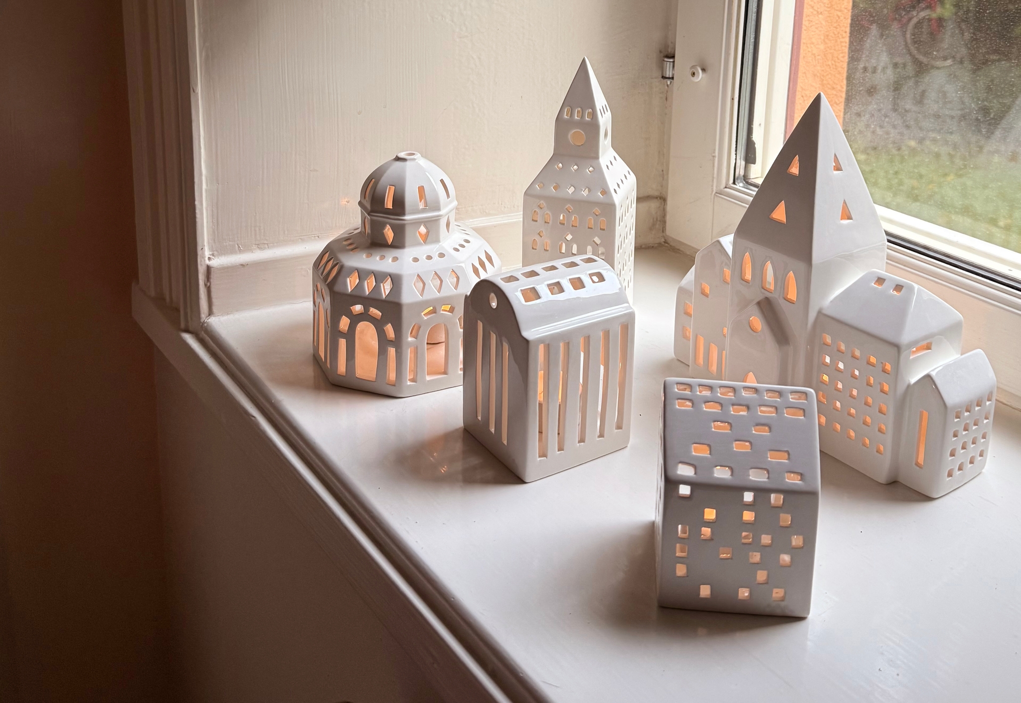 White ceramic miniature buildings and lanterns with glowing lights on a windowsill, beneath dried eucalyptus and red leaves.
