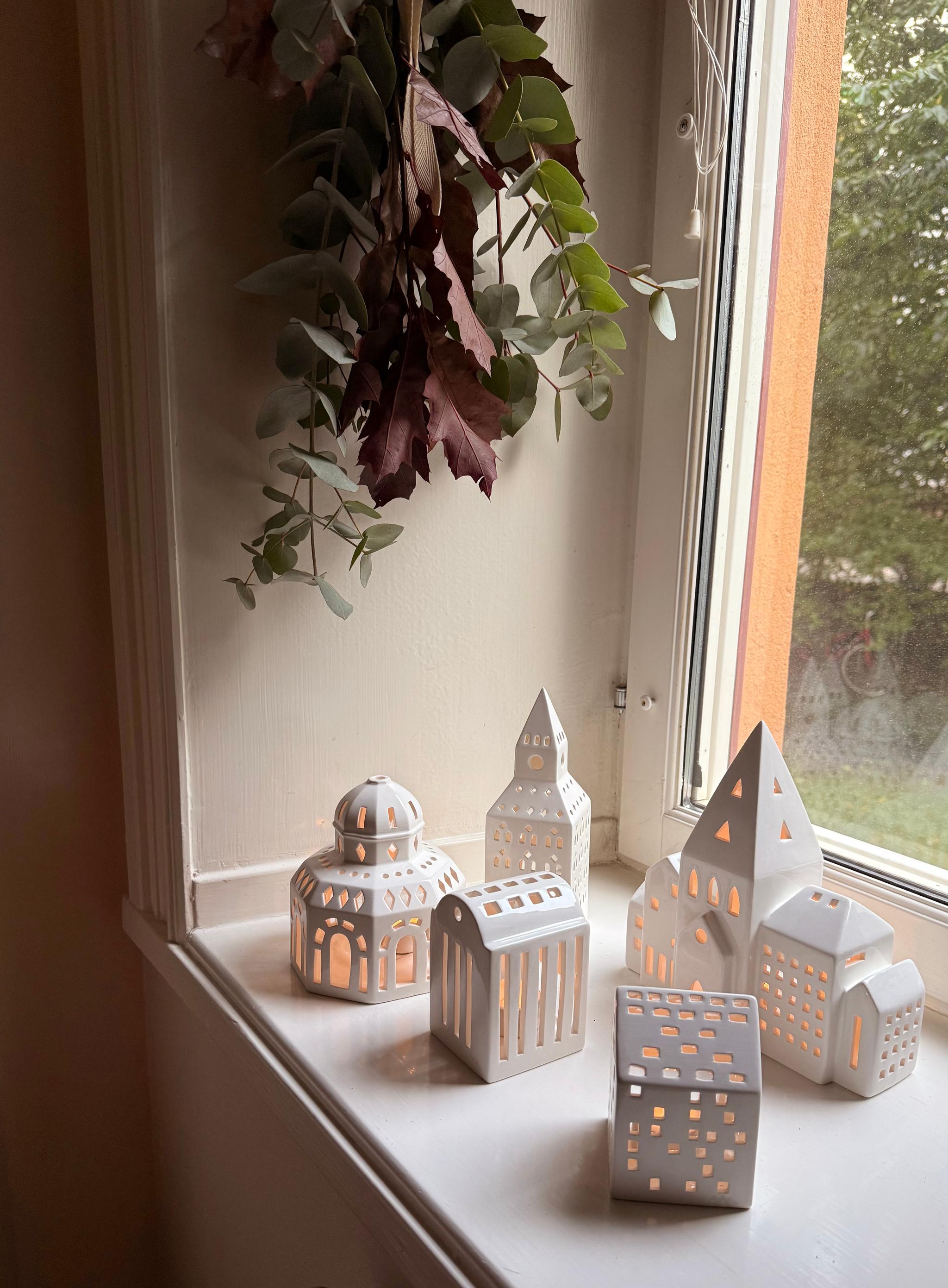 Lit white ceramic house-shaped candle holders on a window sill, beneath a garland of eucalyptus and reddish leaves.