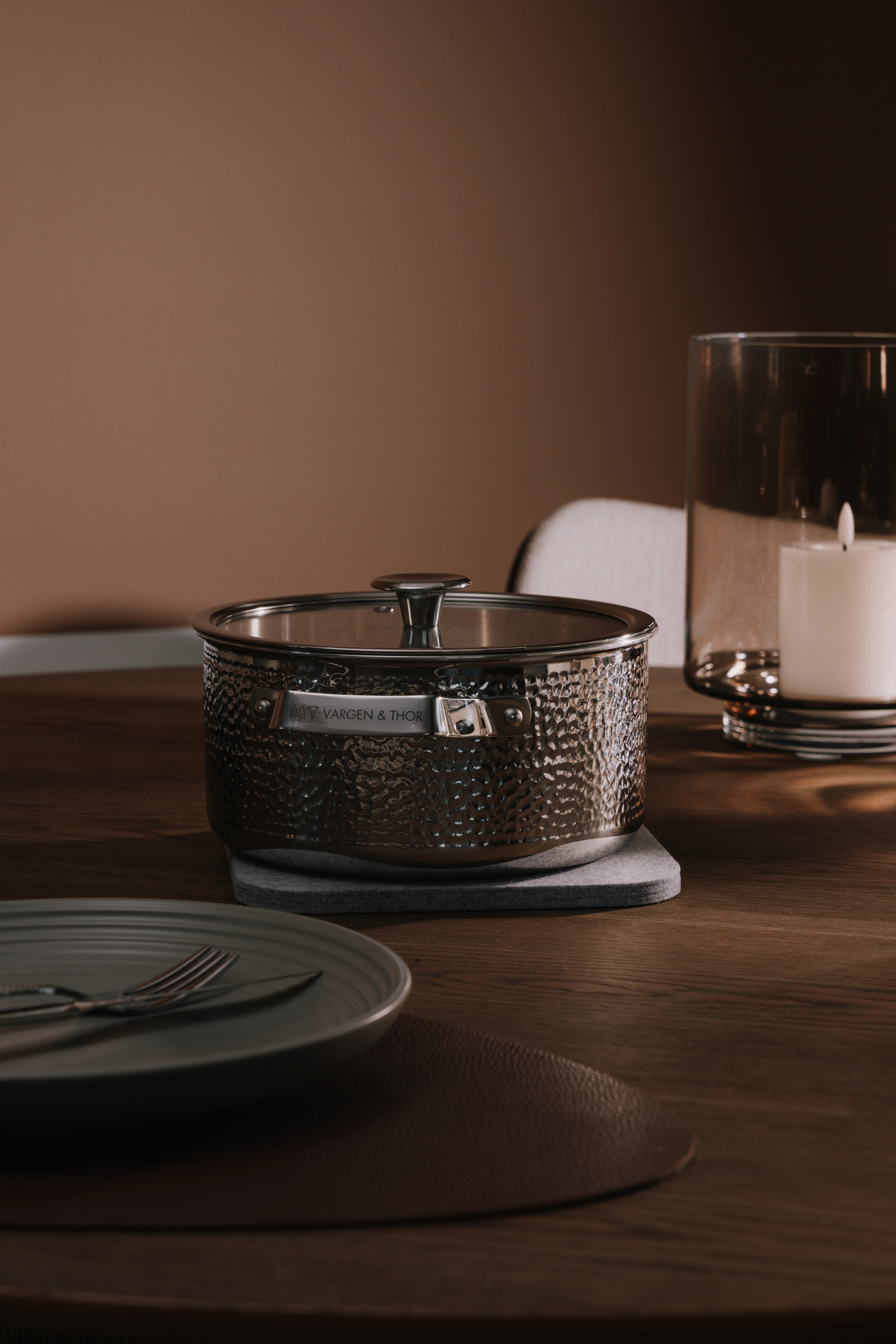 Vasaroitu metallinen Vargen & Thor -kattila lasikannella puupöydällä, lautasen, aterimien ja palavan kynttilän vieressä.