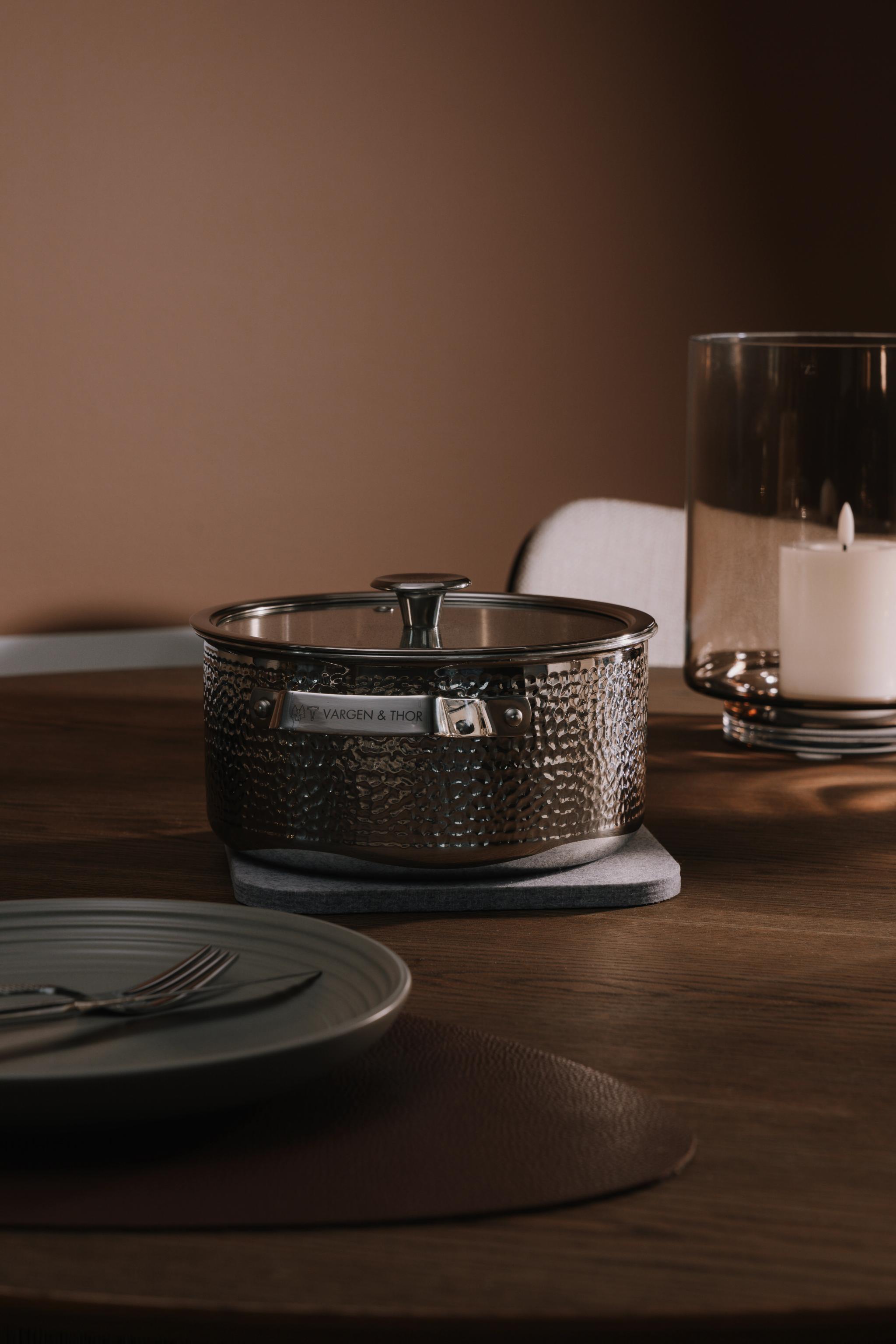 Vasaroitu metallinen Vargen & Thor -kattila lasikannella puupöydällä, lautasen, aterimien ja palavan kynttilän vieressä.