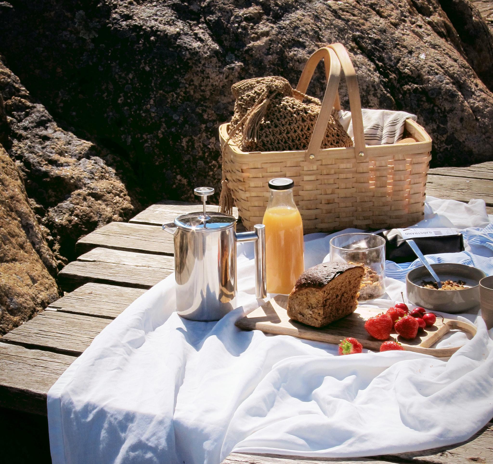Somrig picknick med kaffe, juice, bröd och färska jordgubbar uppdukad på en brygga vid klipporna.