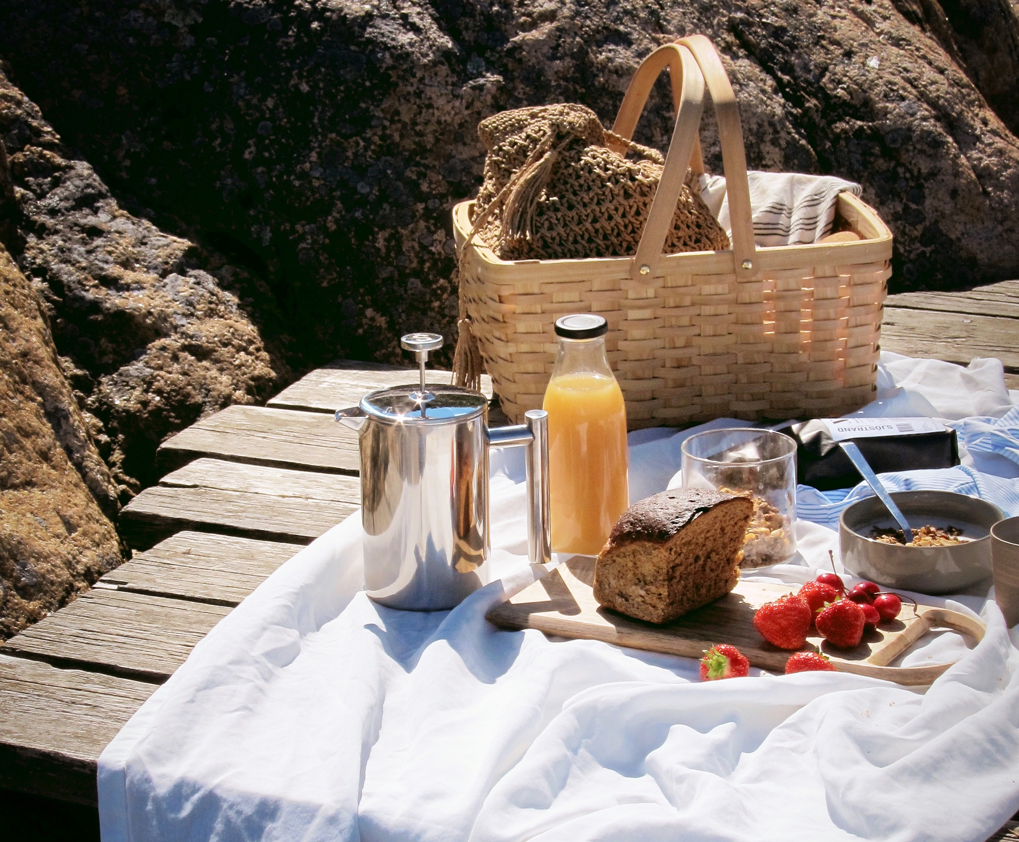 Sommerpiknik på en brygge med kurv, kaffe, juice, brød og friske jordbær foran svabergene.