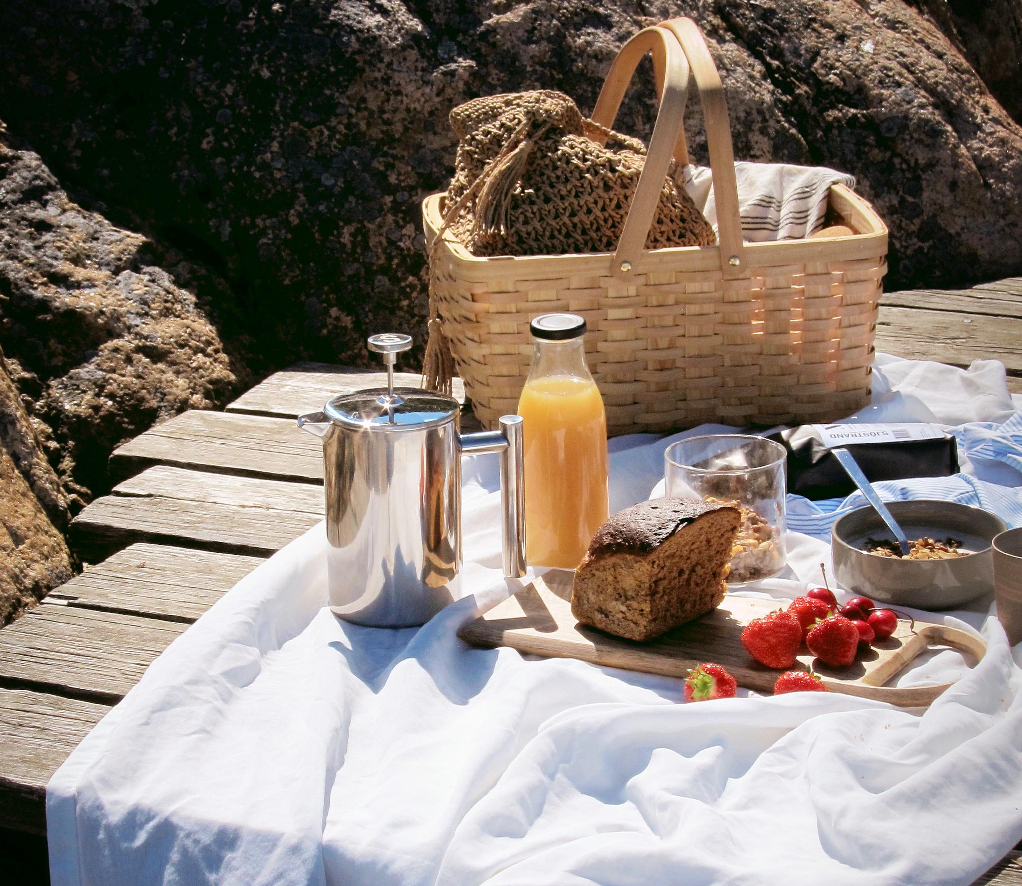 Sommerpiknik ved sjøen med kurv, kaffe, juice, brød og bær på et trebord mot svaberg.