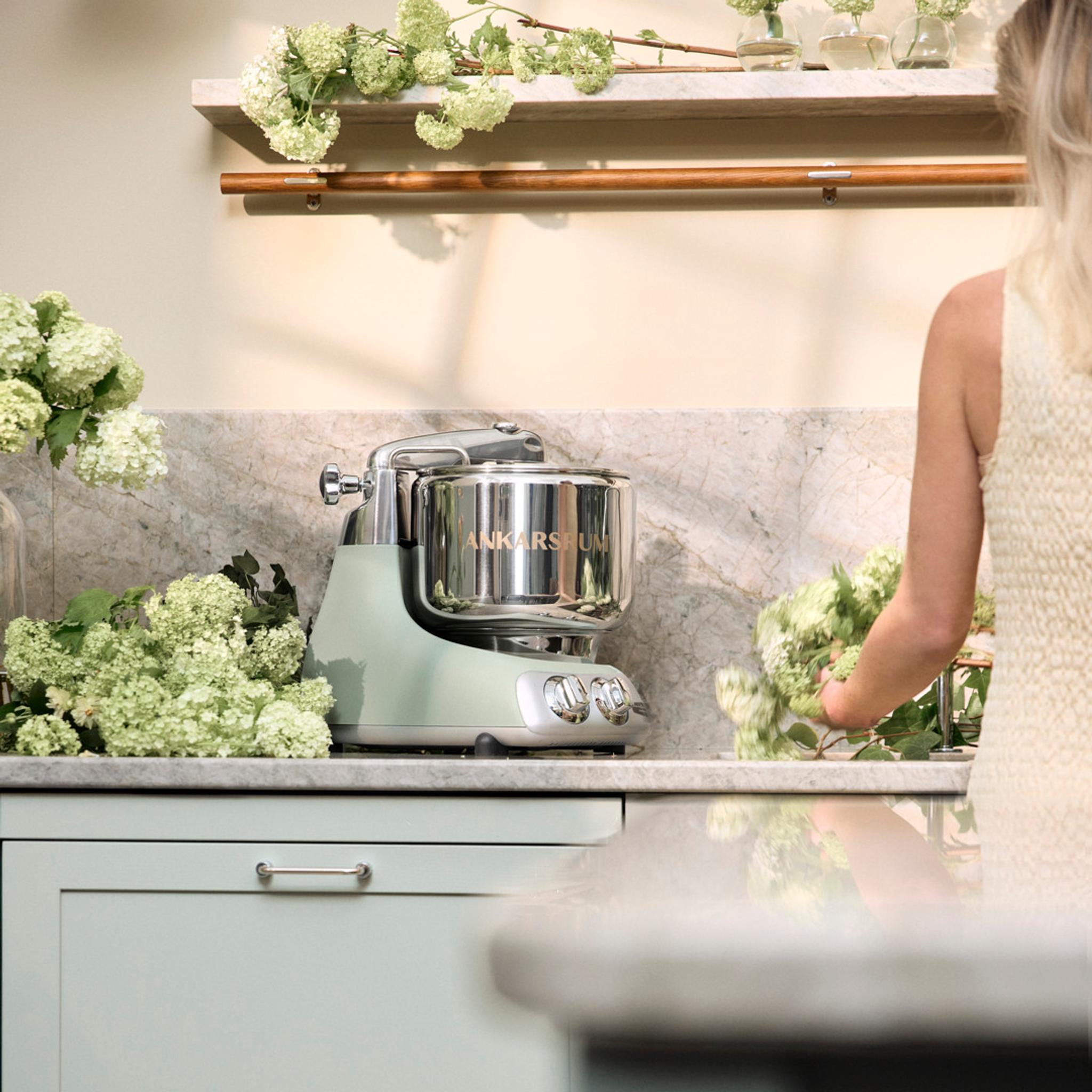 Ankarsrum köksmaskin i pastellgrönt står på en marmorbänk i ett ljust kök medan en person arrangerar gröna blommor bredvid.