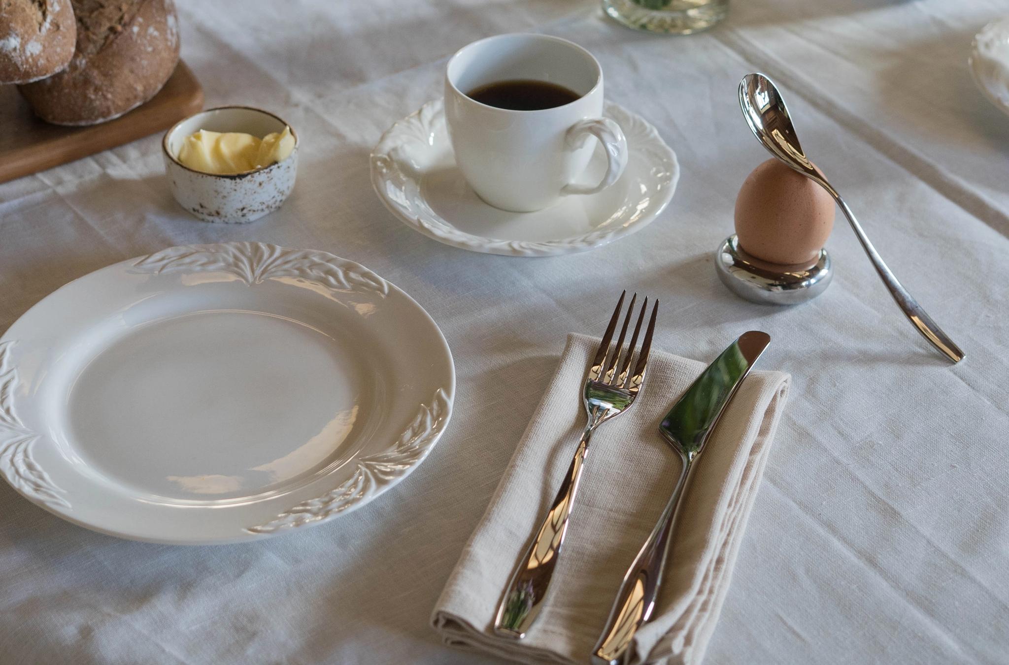 A breakfast table set with a white tablecloth, coffee cups, eggs in holders, bread rolls, butter, and a vase of flowers.
