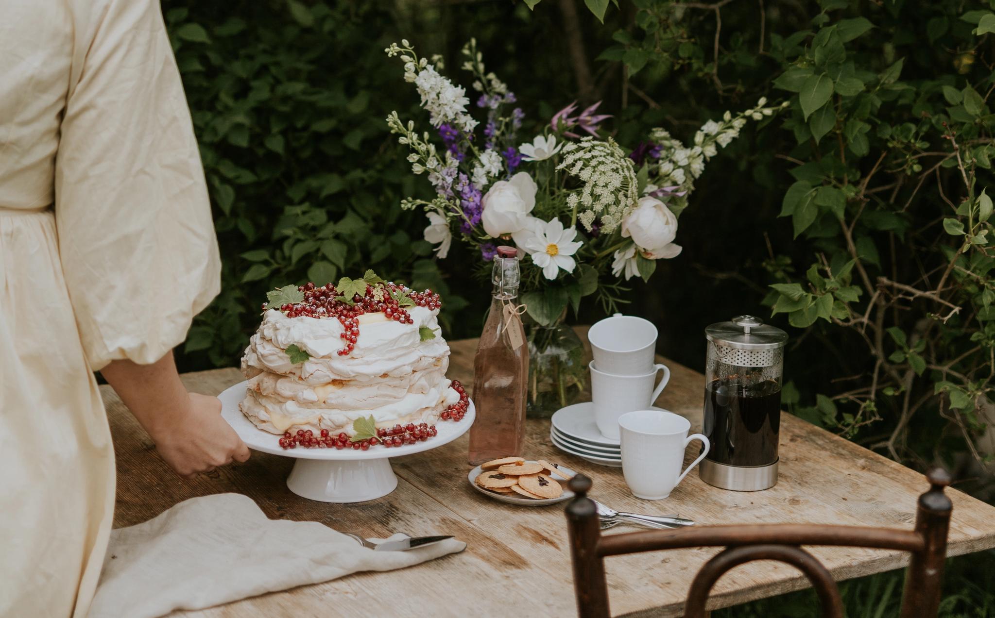 Kvinne dekker et rustikt hagebord med kake, kaffe og blomster foran tett grønn hekk.