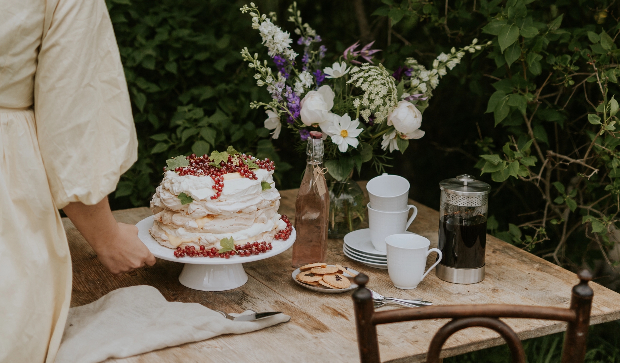 Kvinne dekker et rustikt hagebord med kake, kaffe og blomster foran tett grønn hekk.
