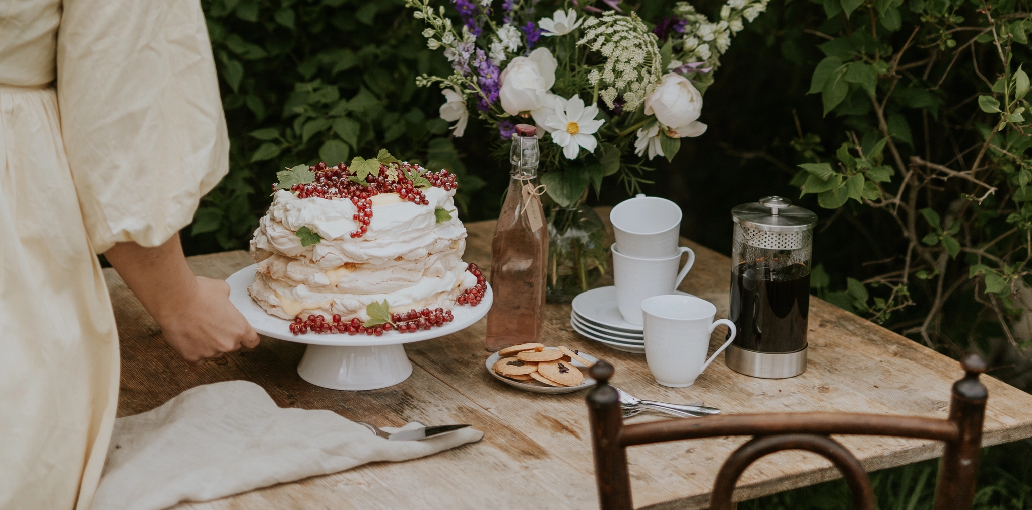 Kvinna dukar ett rustikt trädgårdsbord med sommartårta, kaffe och blommor framför lummig grönska.