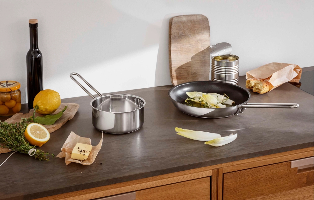 Keittiötaso, jolle on aseteltu tuoreita raaka-aineita, kuten sitruunoita, yrttejä, voita, oliiveja ja leipää, kattilan ja sikurilla täytetyn paistinpannun vieressä