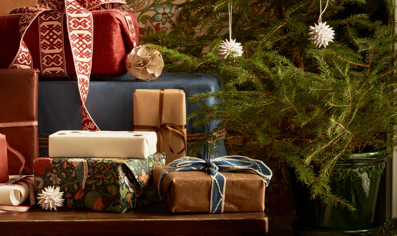 A small Christmas tree decorated with white paper star ornaments next to wrapped presents, against floral wallpaper and a large red paper star.