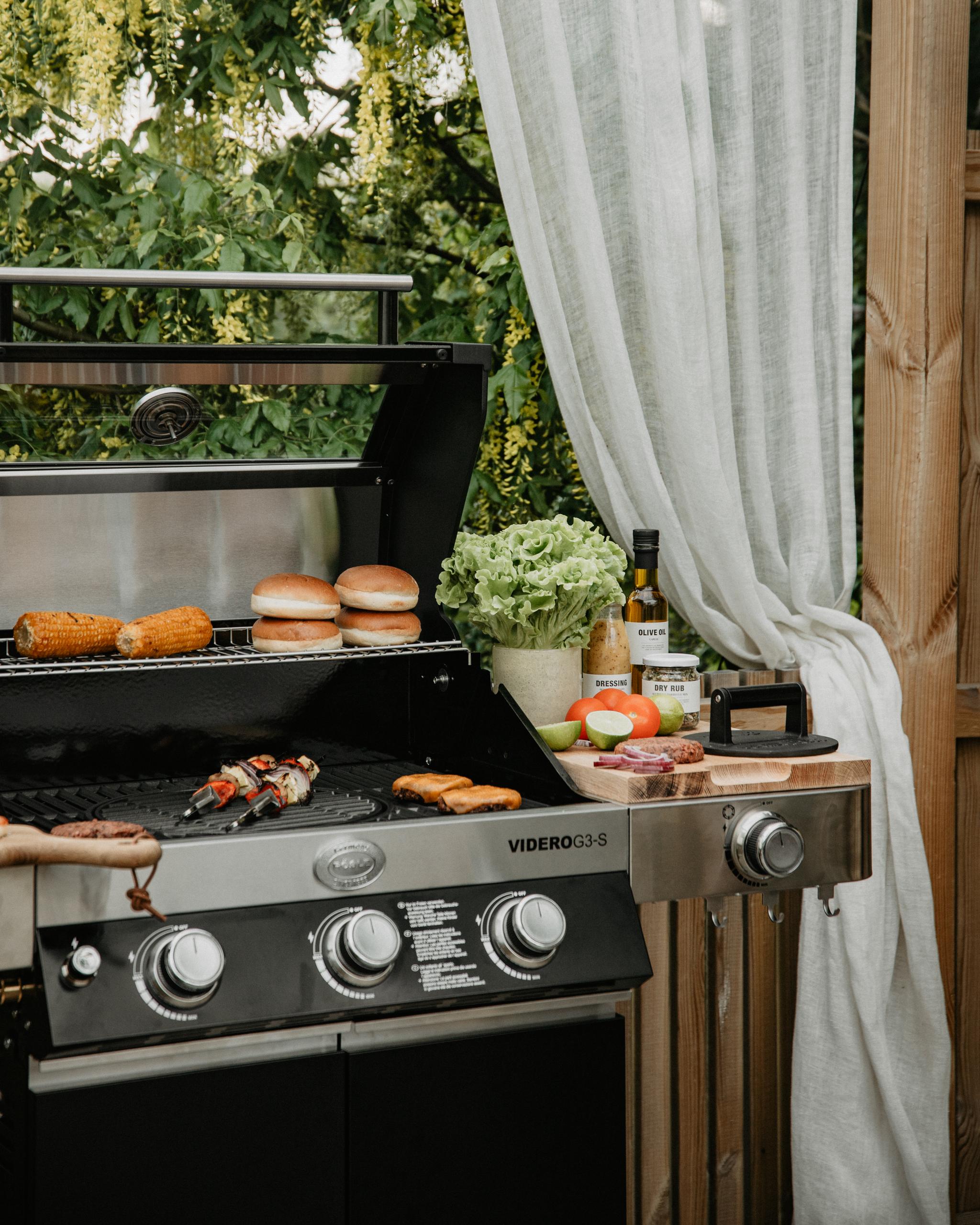 En gasolgrill utomhus där majs, bröd och kebab tillagas, med hamburgare och färska grönsaker på en skärbräda