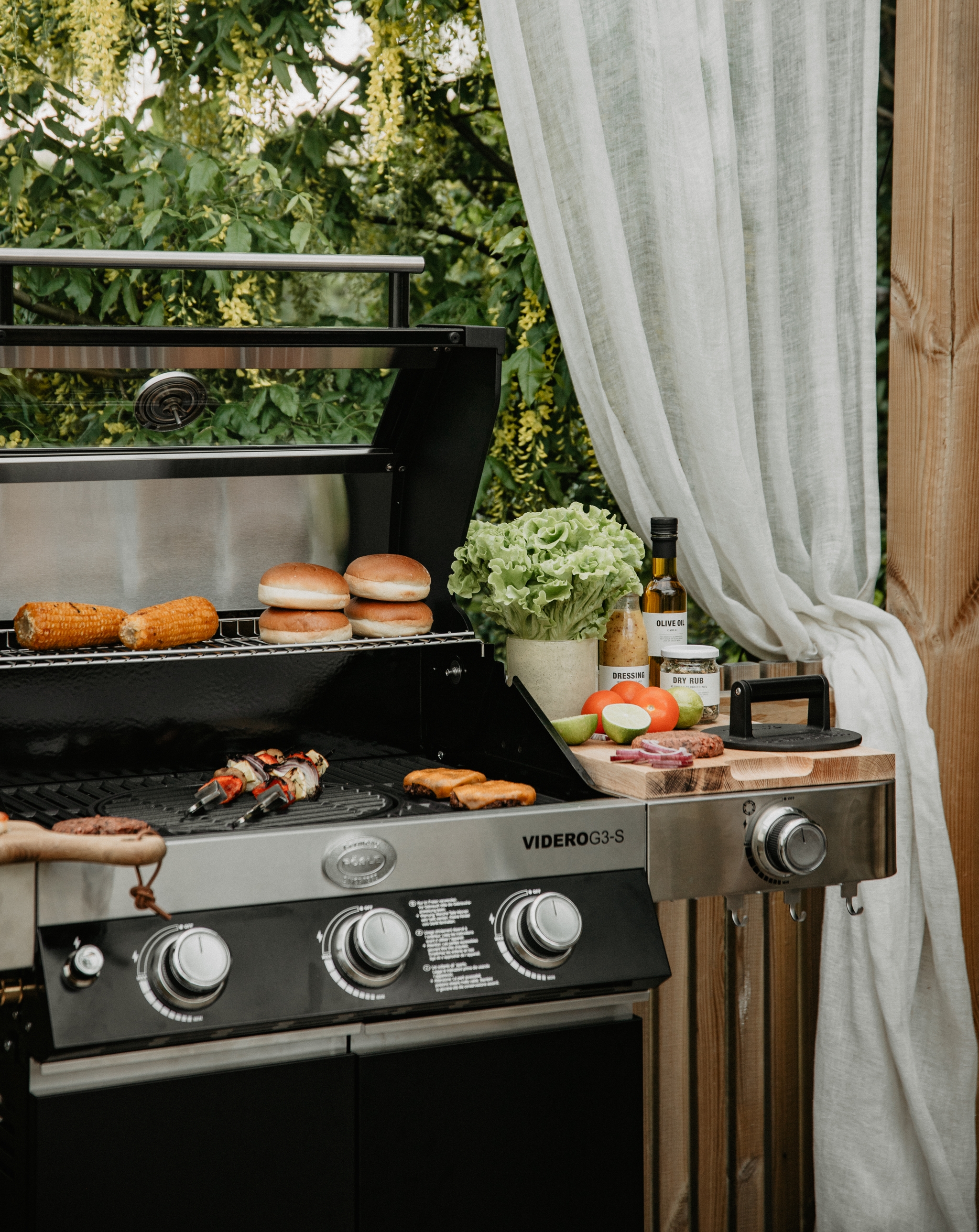 En gasolgrill utomhus där majs, bröd och kebab tillagas, med hamburgare och färska grönsaker på en skärbräda