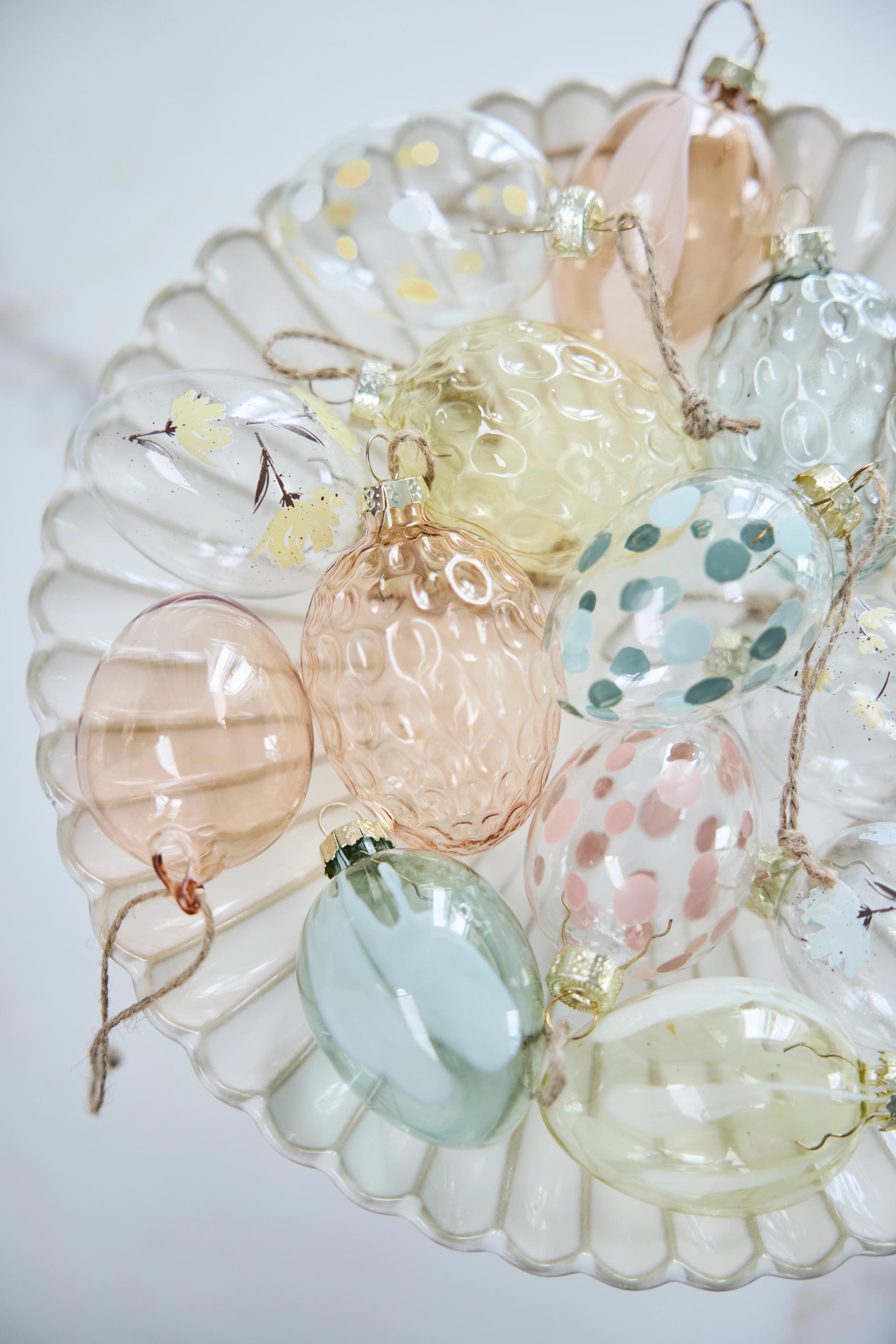 Assorterte eggepynt i pastellfarget glass, noen klare med mønstre, på et hvitt, kamskjellformet fat.