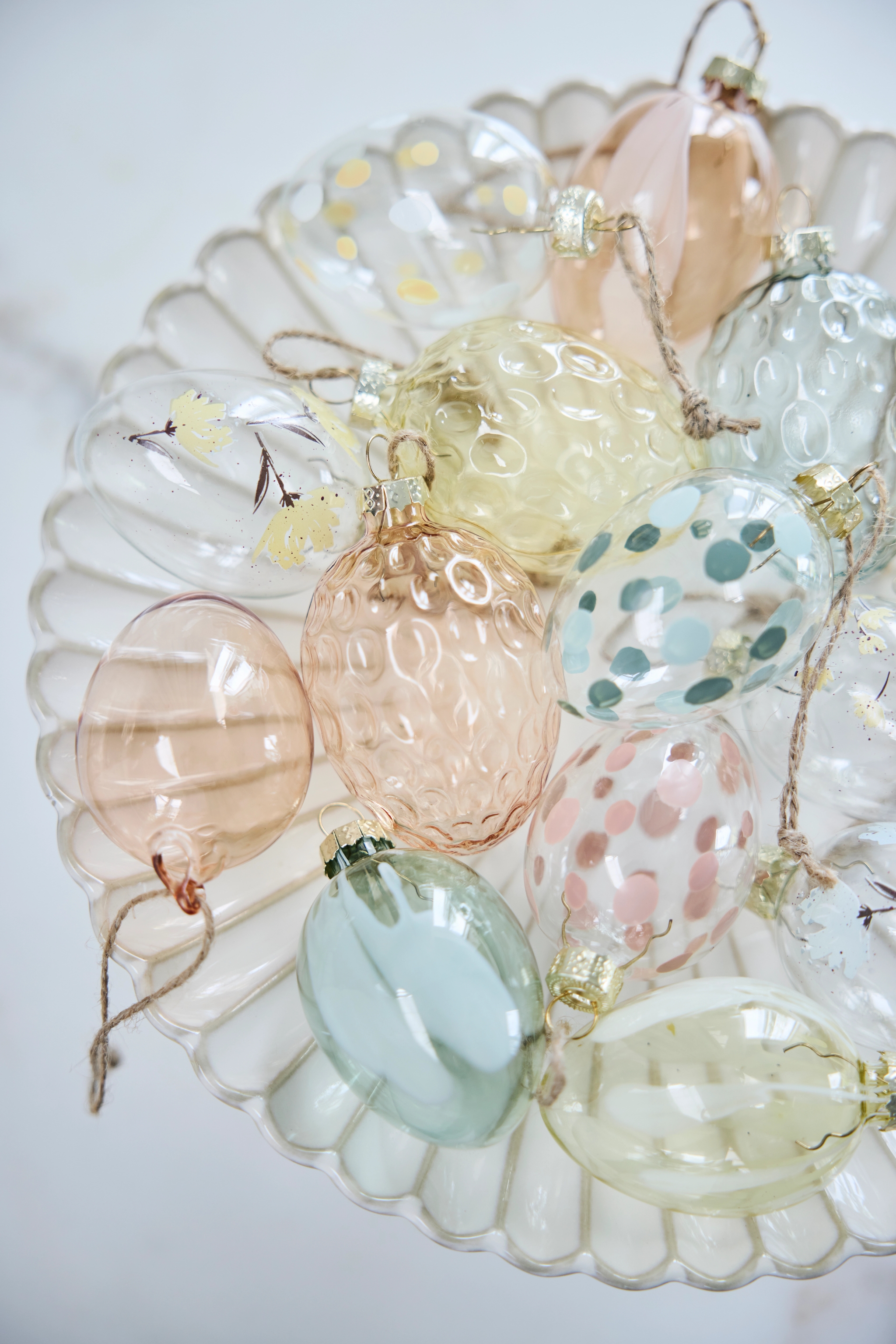 Assorterte eggepynt i pastellfarget glass, noen klare med mønstre, på et hvitt, kamskjellformet fat.
