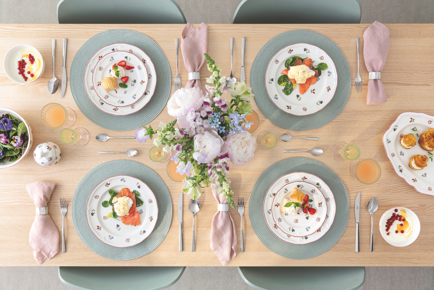 Vakkert dekket spisebord sett ovenfra med blomstrete porselen, desserter, juicedrikker og en stor bukett pastellfargede blomster i midten.