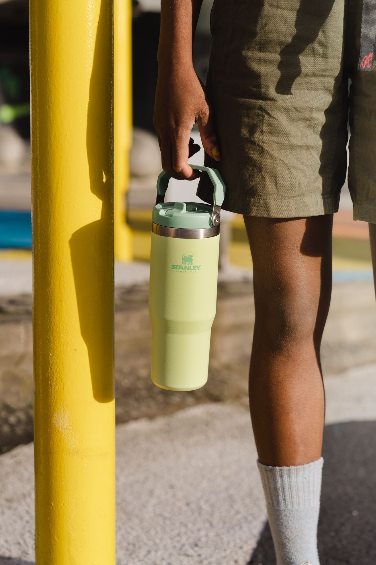A person holds a pale green Stanley tumbler with a handle, standing next to a bright yellow pole.