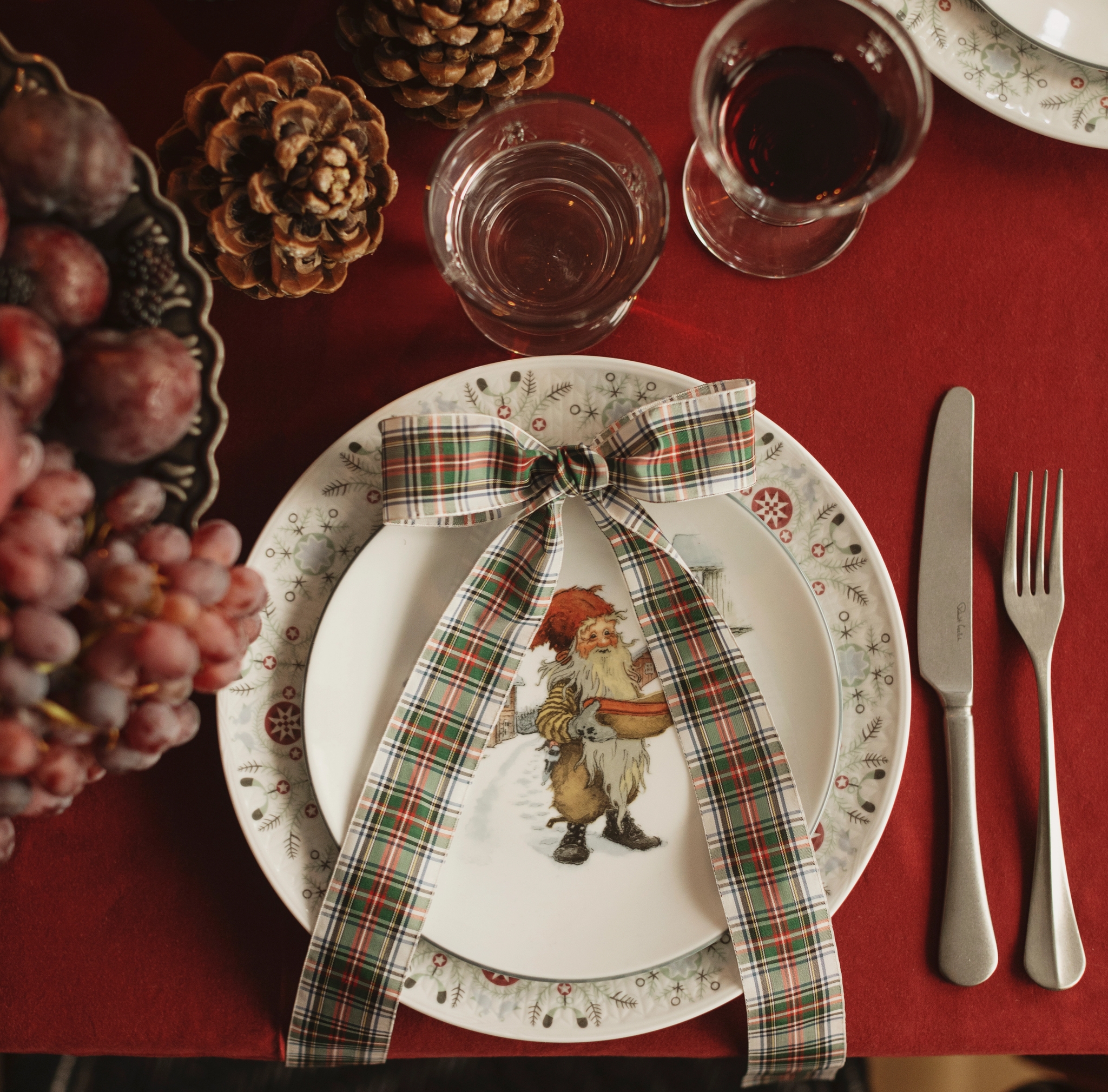 En juldukning med en röd duk, tallrikar med en jultomteillustration och en rutig rosett, bestick, drycker, kottar och vindruvor.