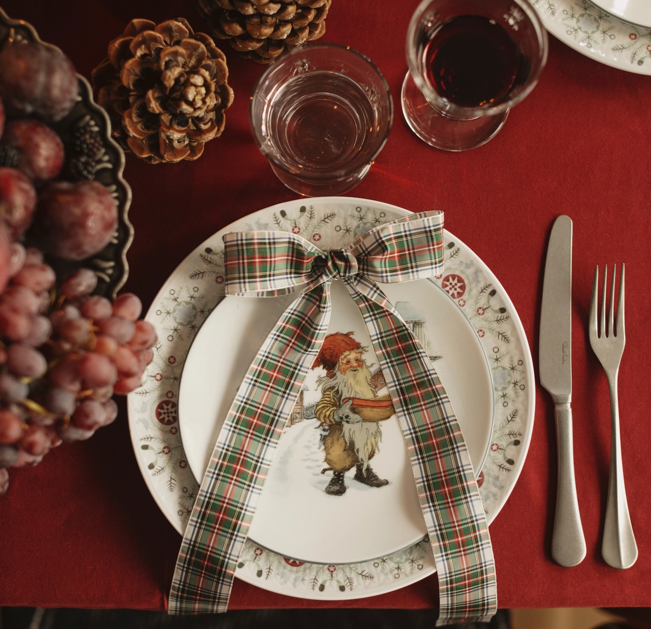 En festlig juldukning med en röd duk, en tallrik med jultomten, rutigt band, tallkottar, vindruvor och glas vin och vatten.