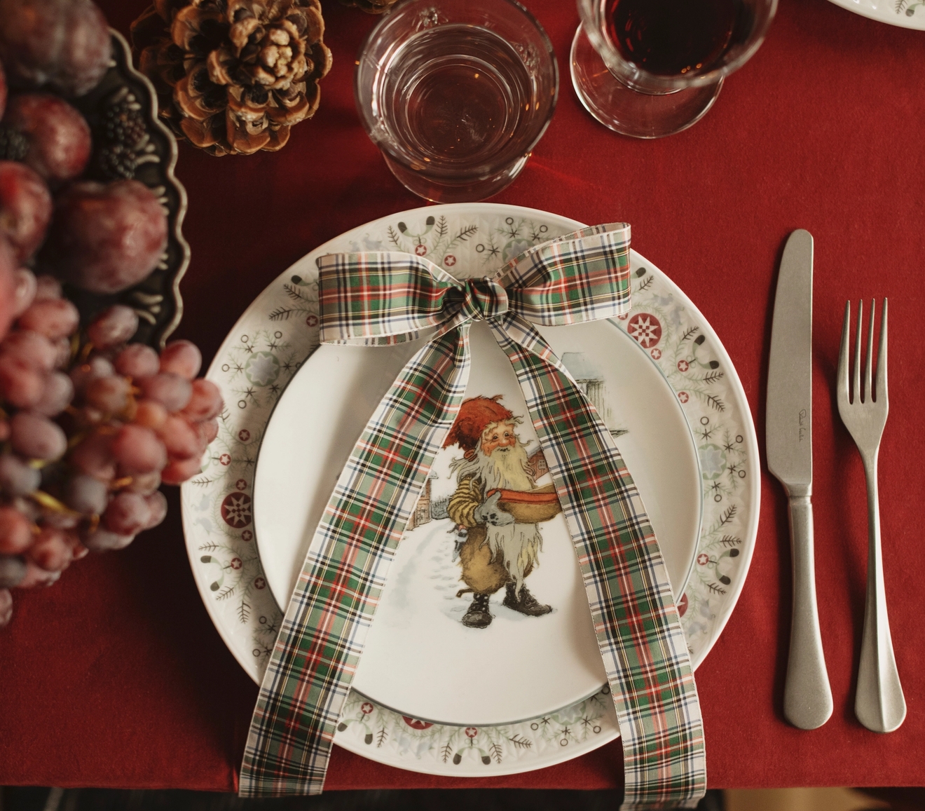 En festlig juldukning med en röd duk, en tallrik med jultomten, rutigt band, tallkottar, vindruvor och glas vin och vatten.