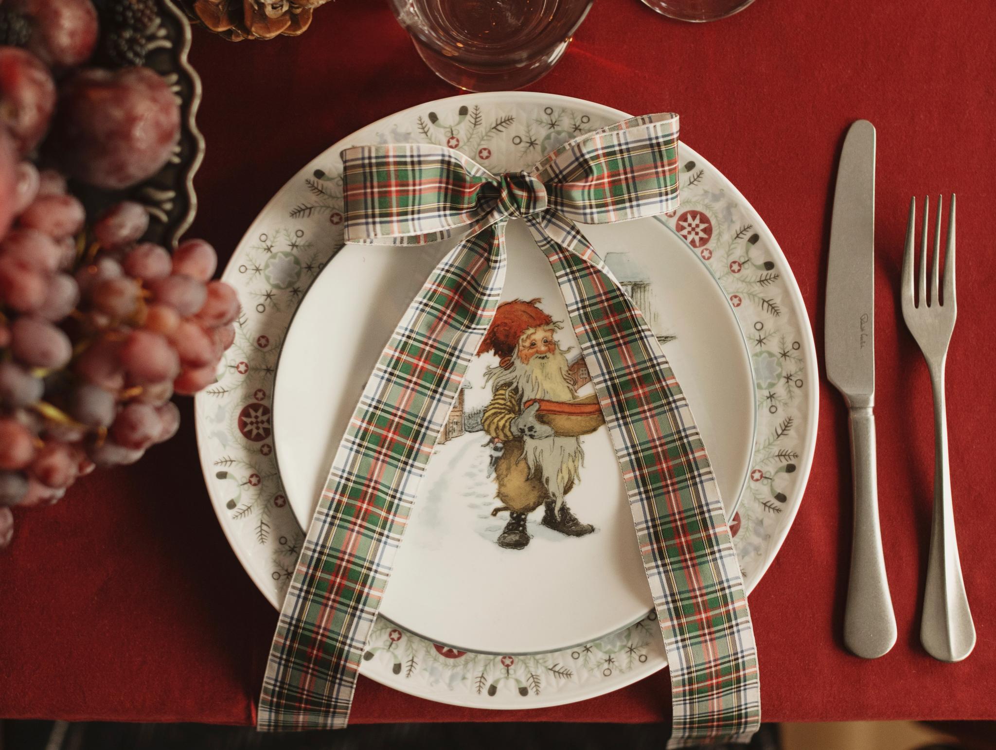 En juldukning med en röd duk, tallrikar med en jultomteillustration och en rutig rosett, bestick, drycker, kottar och vindruvor.