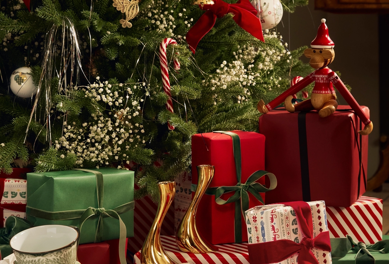 Joulukuusi, joka on koristeltu kimaltavalla nauhalla, koristeilla ja valkoisilla kukilla. Sen juurella on punaisia lahjapaketteja sekä puinen tonttuhahmo, joka istuu yhden paketin päällä.