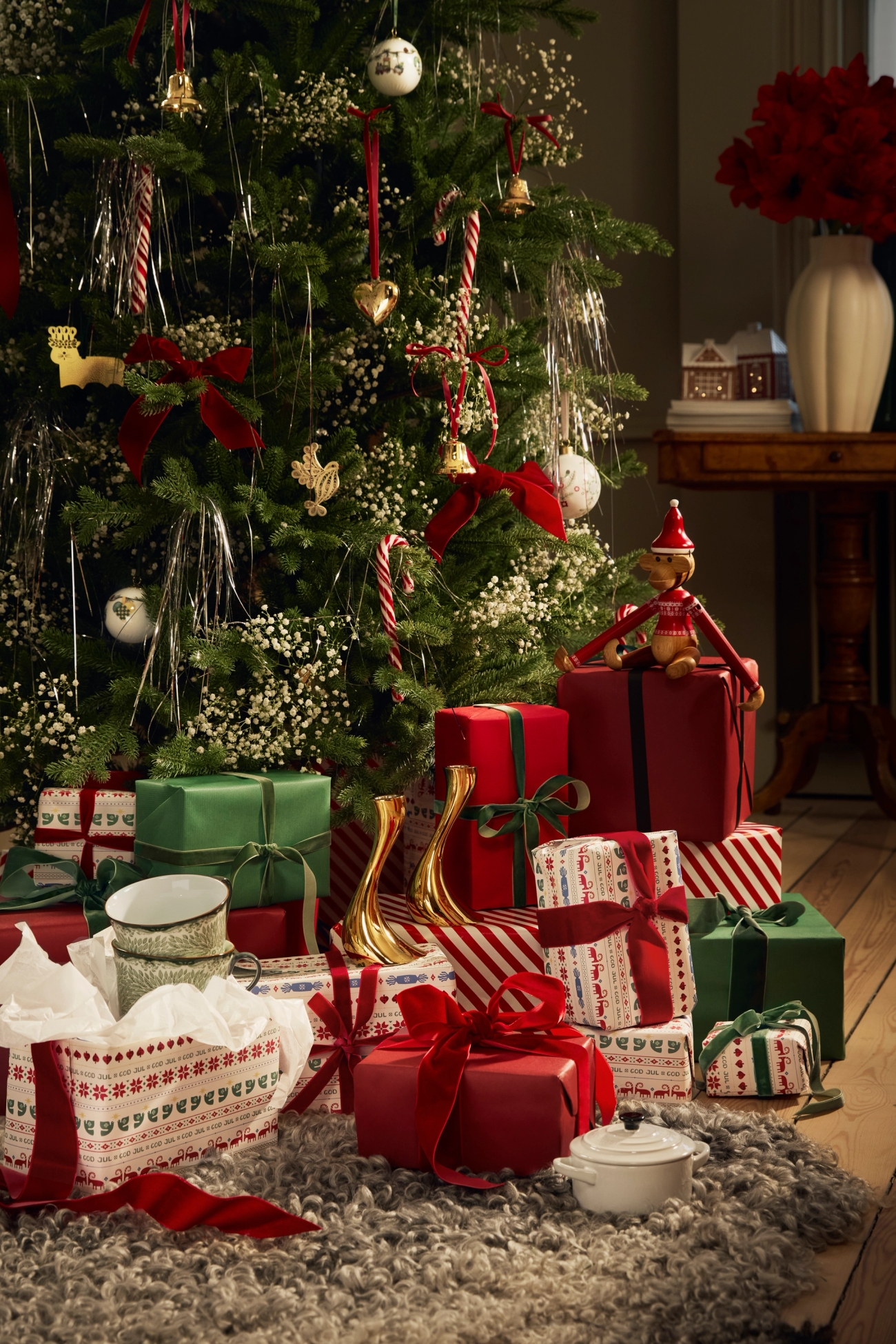 A festive scene with a decorated Christmas tree, many red and green wrapped presents on a gray shag rug, an elf doll, and a table with red flowers.