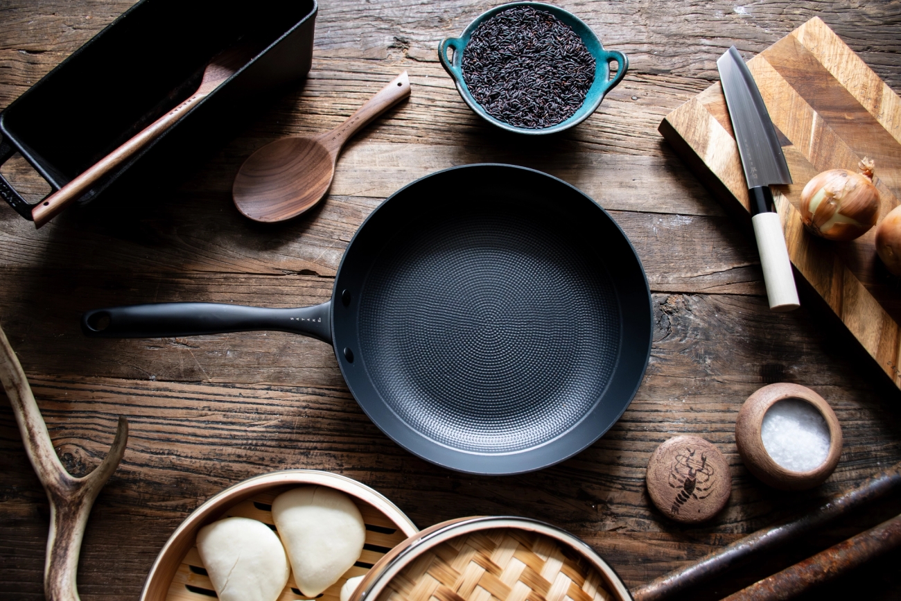 En mörk stekpanna från Satake, svart ris, lök och salt är arrangerade med matlagningsredskap på ett rustikt träbord.