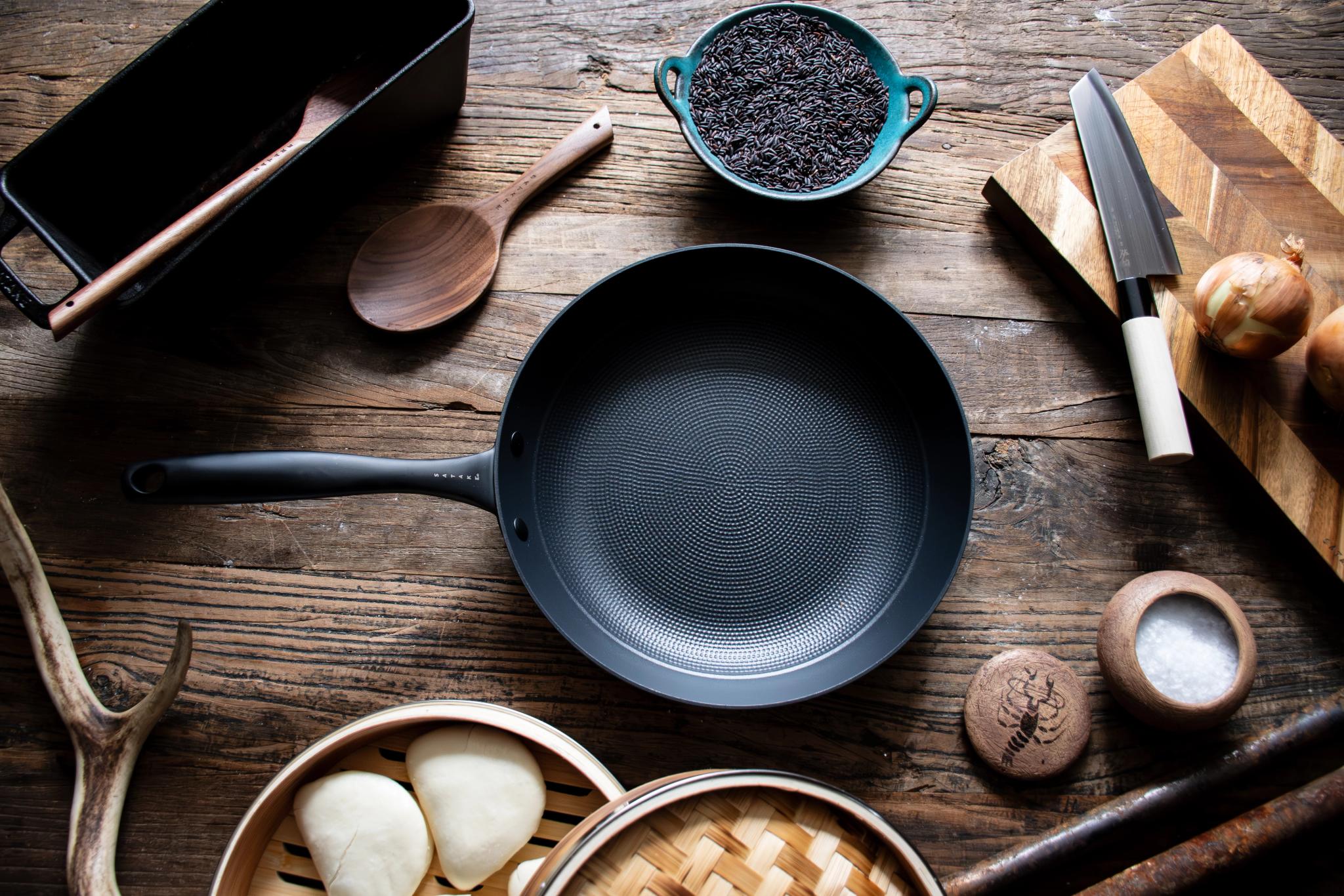 Bild ovanifrån av en mörk stekpanna, matlagningsverktyg och ingredienser på ett rustikt träbord.