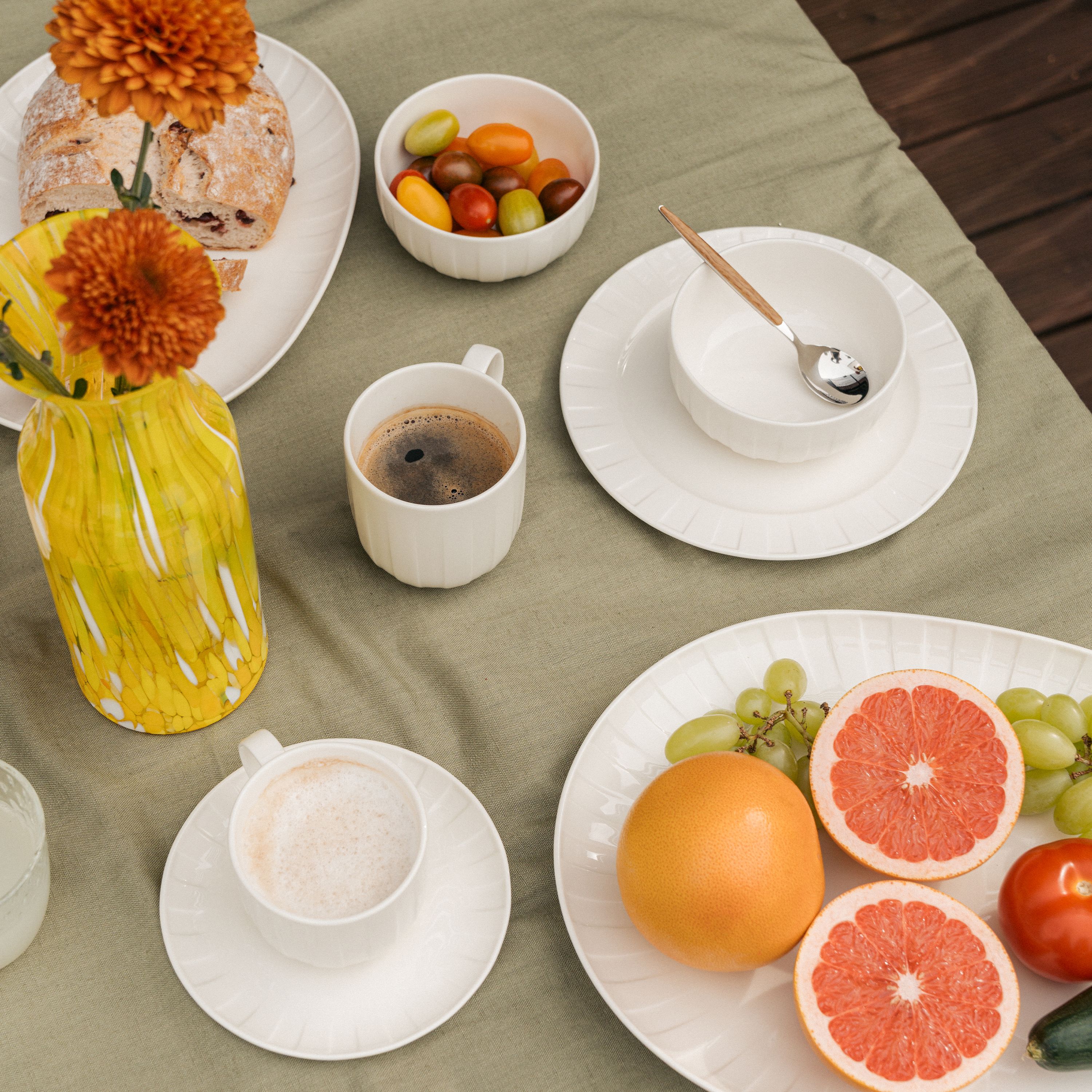 Sommerlig frokostbord ute med kaffe, fersk grapefrukt, druer, brød og fargerike tomater på hvitt servise.