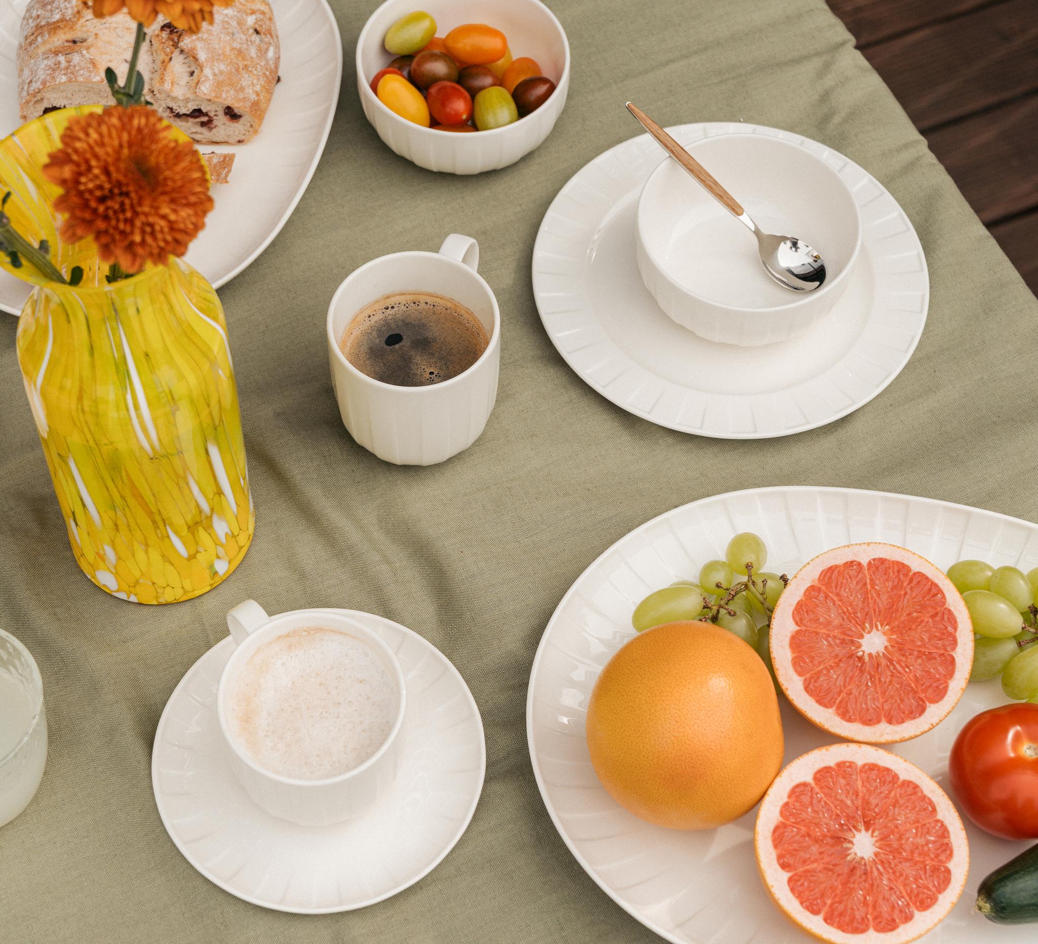 Sommerlig frokostbord ute med kaffe, fersk grapefrukt, druer, brød og fargerike tomater på hvitt servise.