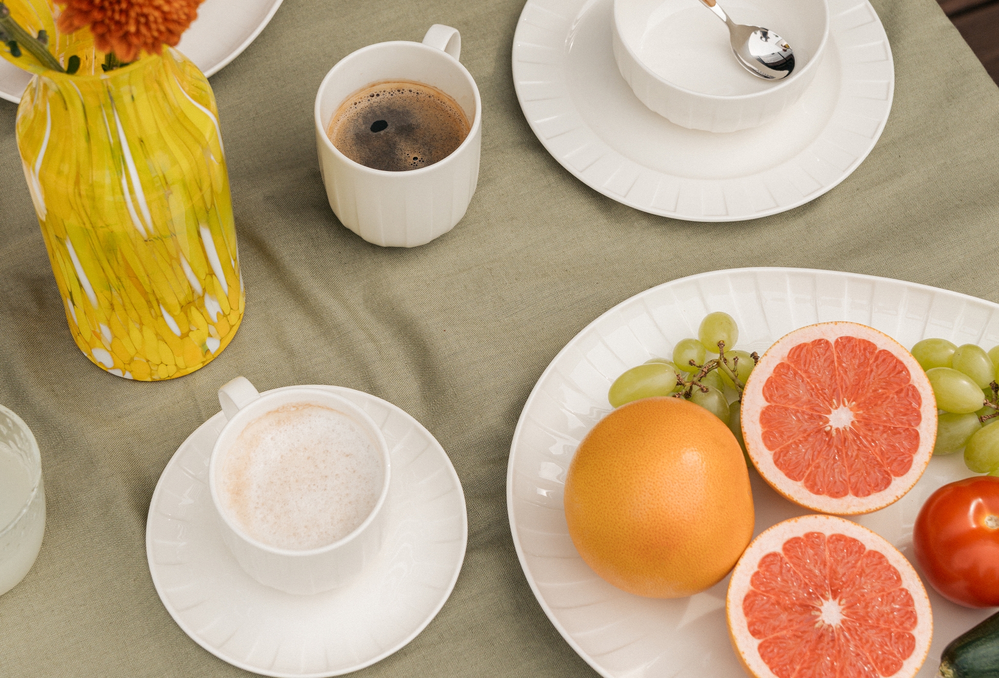 Dekket frokostbord med kaffe, fersk frukt, brød og blomster på grønn duk.