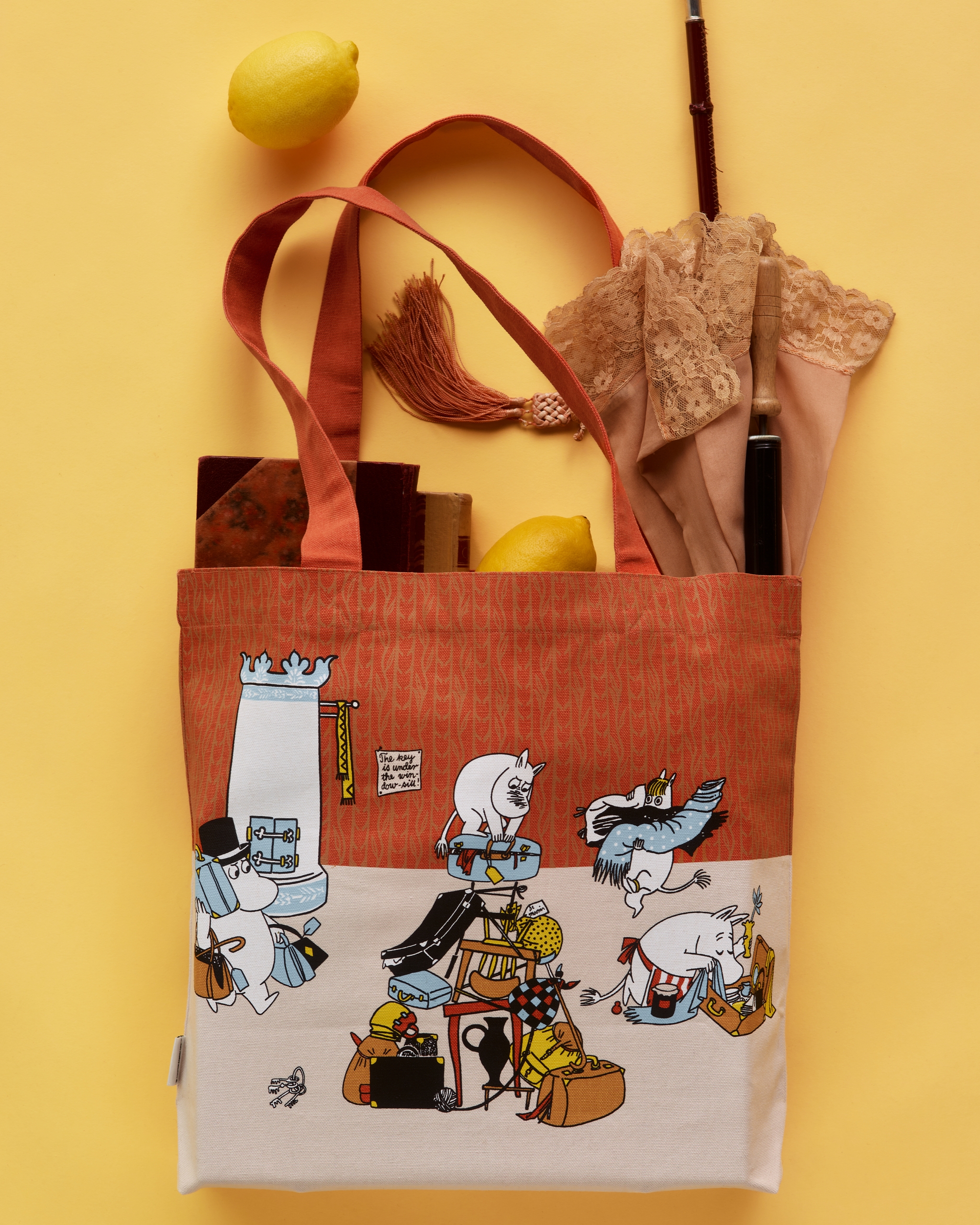 Orange och vit Mumin-tygväska föreställande karaktärer som packar, fylld med böcker, citroner och ett spetsparaply, mot gul bakgrund.