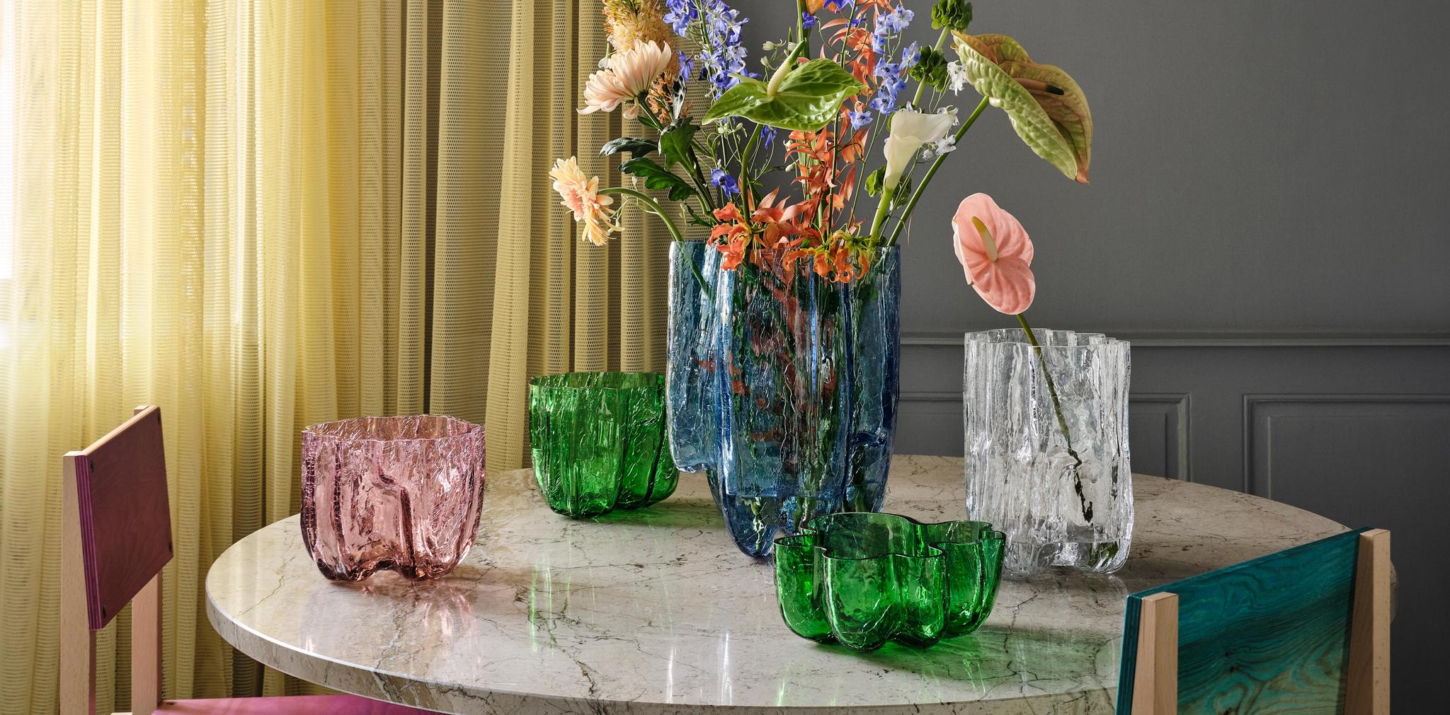 Färgglada glasvaser med blommor står på ett runt marmorbord framför ljusa gardiner i ett modernt vardagsrum.