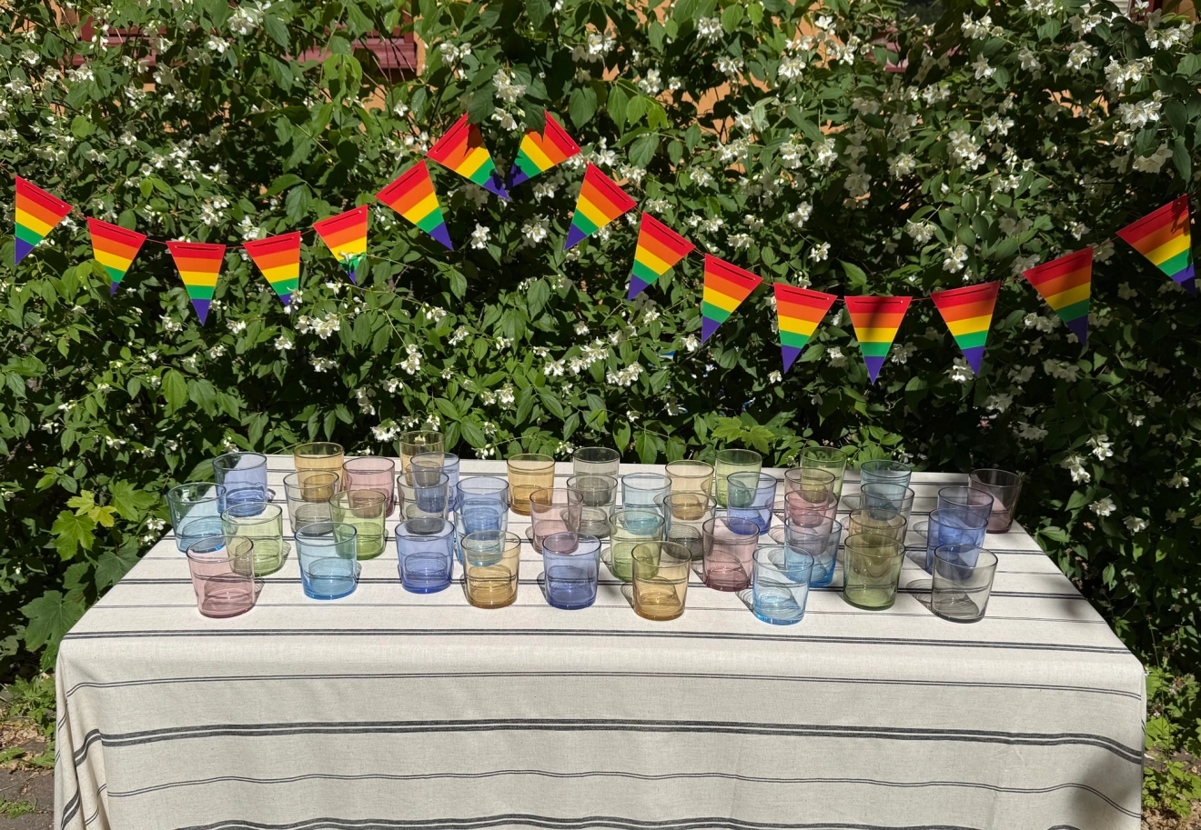 Ett utomhusbord med randig duk är dukat med många färgade glas framför en grön häck dekorerad med regnbågsflaggor för Pride.