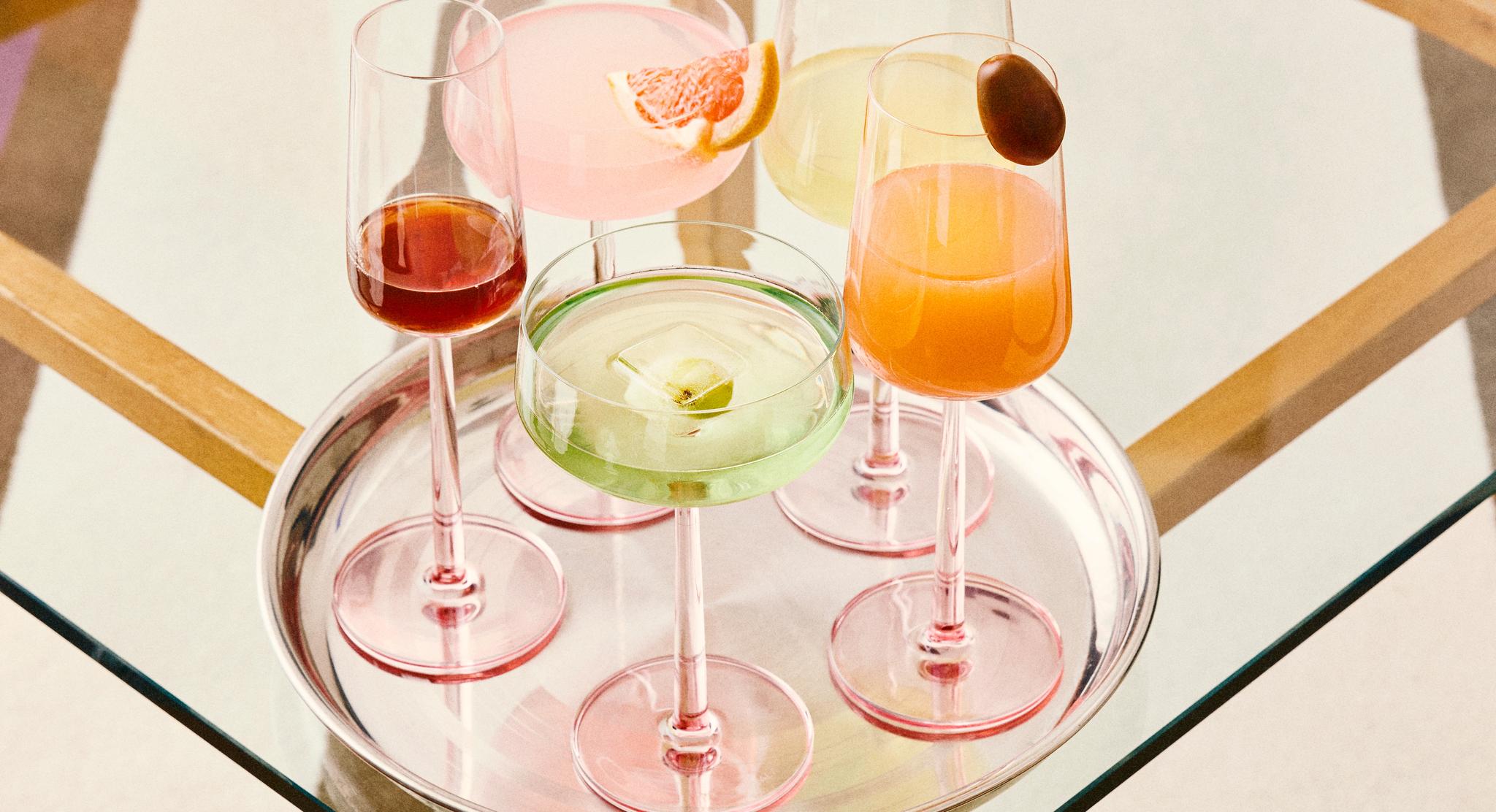 Neljä vaaleanpunaisilla jaloilla varustettua lasia ja värikkäitä juomia hopeisella tarjottimella lasipöydällä.