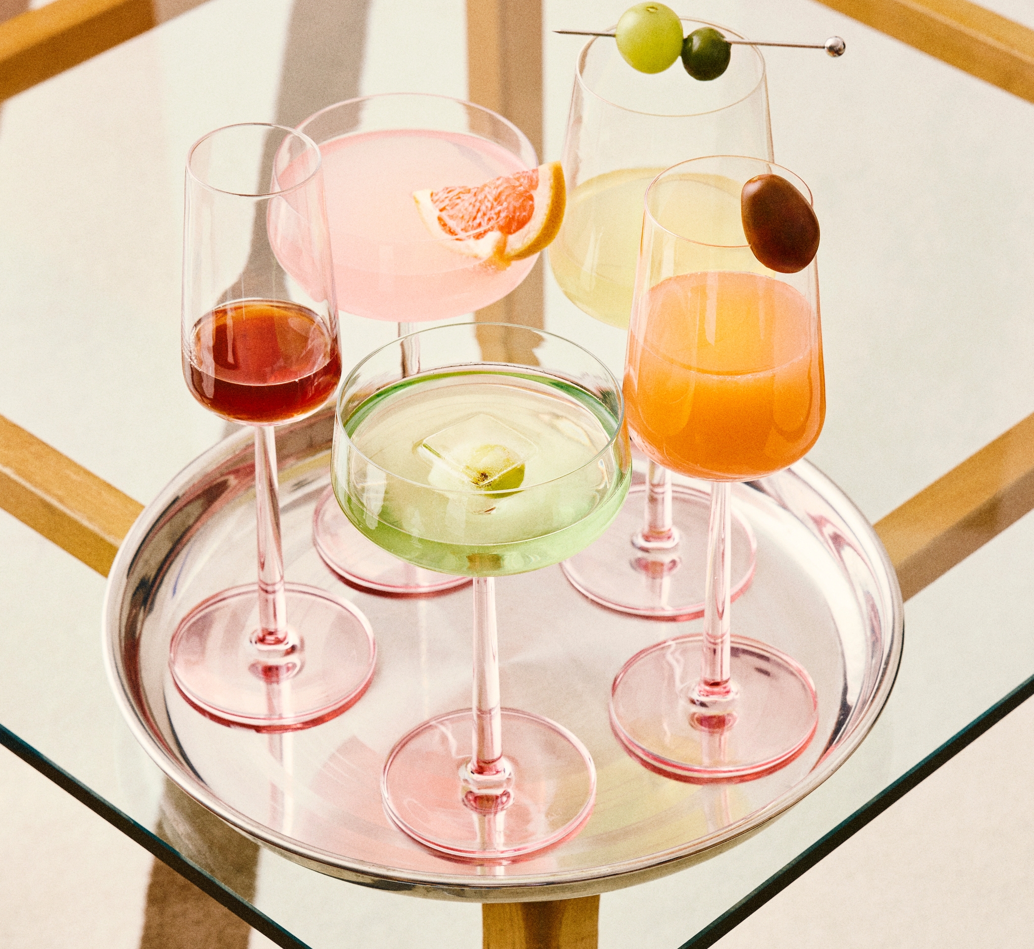 Neljä vaaleanpunaisilla jaloilla varustettua lasia ja värikkäitä juomia hopeisella tarjottimella lasipöydällä.