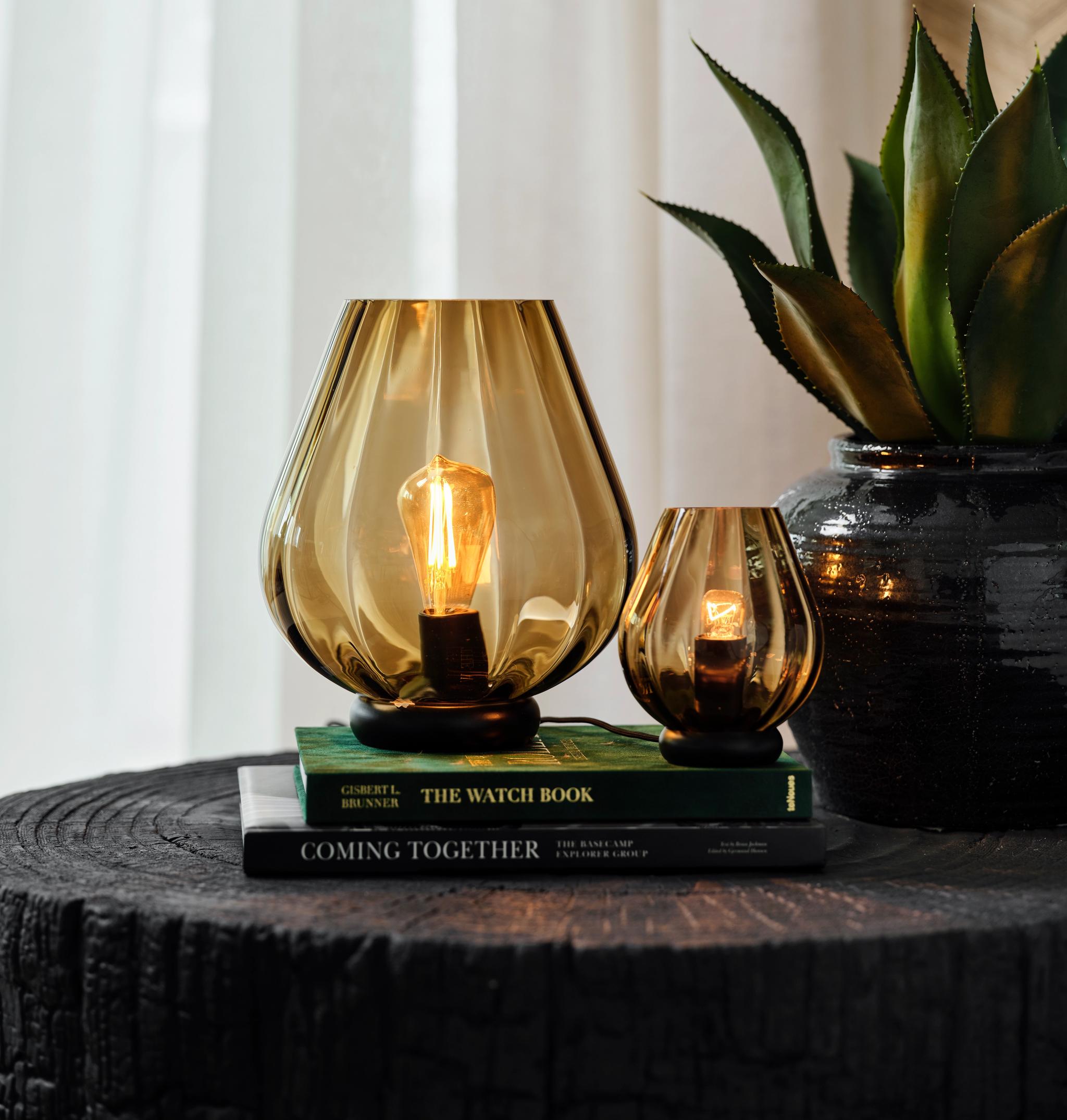 To dekorative bordlamper i ravfarget glass står på bøker ved siden av en grønn plante.