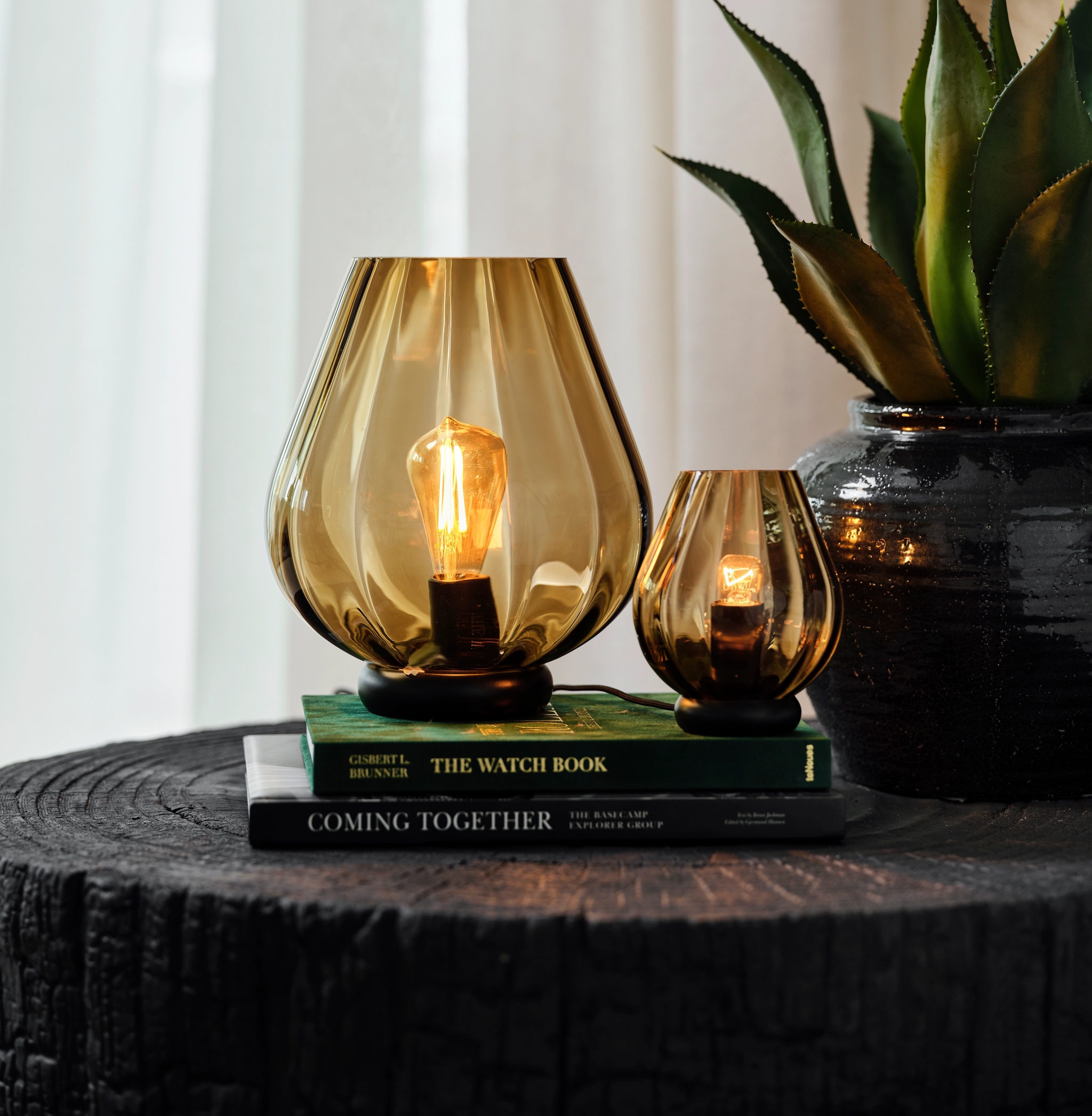 To dekorative bordlamper i ravfarget glass står på bøker ved siden av en grønn plante.