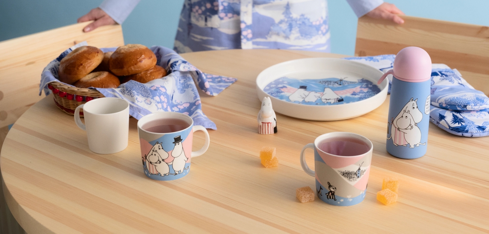a woman is standing at a table with a basket of bread and mugs .