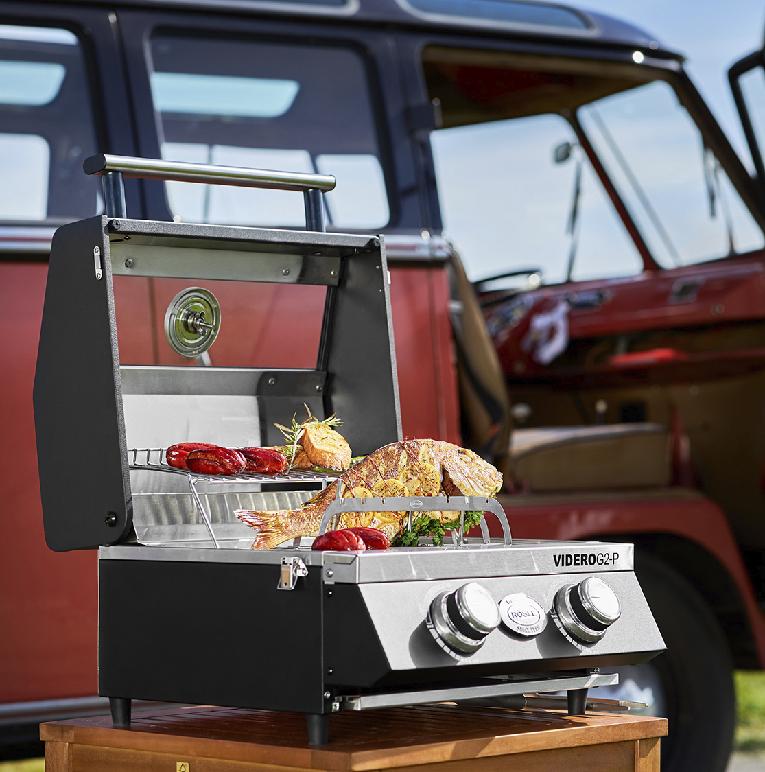A black and stainless steel gas grill with a whole fish and sausages cooking on it, in front of a red vintage vehicle.