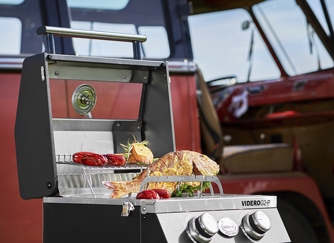 A black and stainless steel gas grill with a whole fish and sausages cooking on it, in front of a red vintage vehicle.