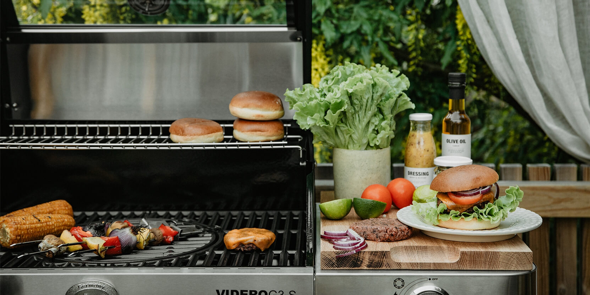 A gas grill cooking corn, vegetable skewers, and a burger patty, with fresh ingredients and a prepared burger on a cutting board nearby.