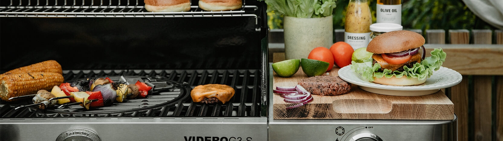 A gas grill cooking corn, vegetable skewers, and a burger patty, with fresh ingredients and a prepared burger on a cutting board nearby.