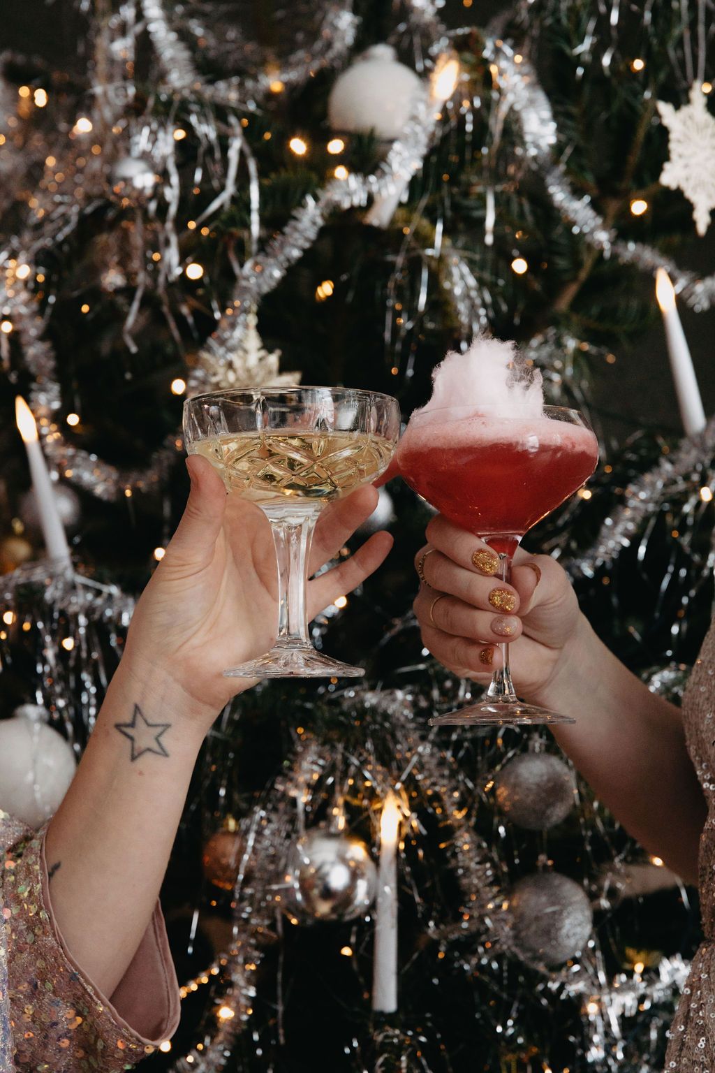 Två händer skålar med ett mousserande vin och en röd cocktail toppad med sockervadd, framför en festlig julgran.
