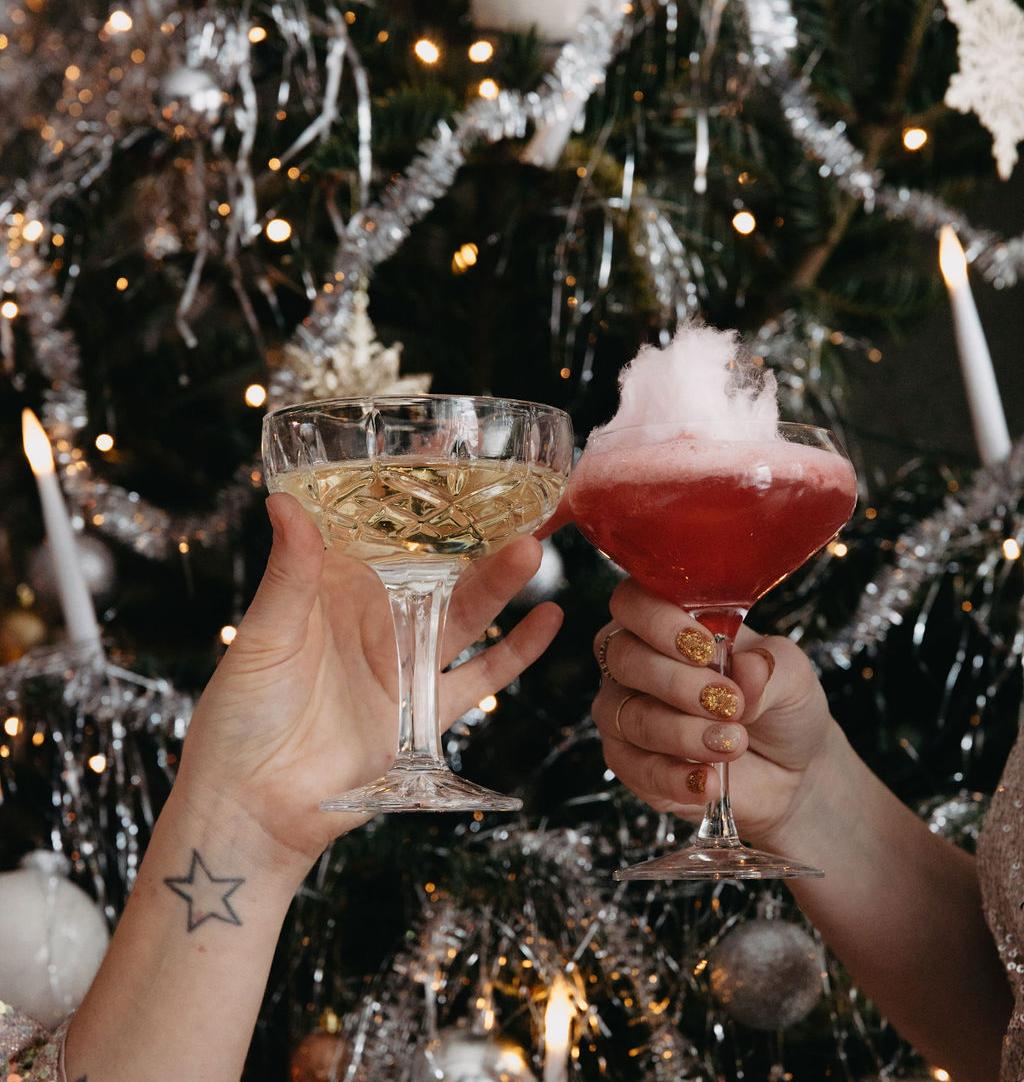 Två händer skålar med ett mousserande vin och en röd cocktail toppad med sockervadd, framför en festlig julgran.