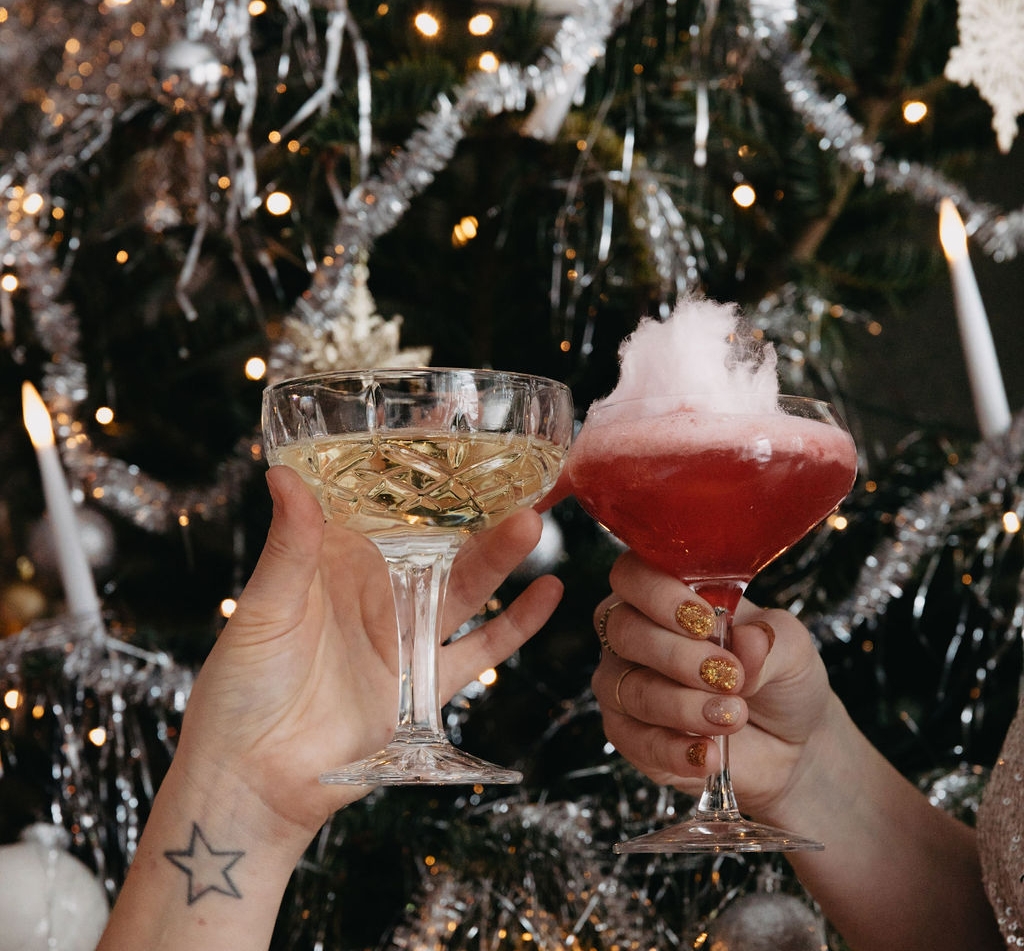 Två händer skålar med ett mousserande vin och en röd cocktail toppad med sockervadd, framför en festlig julgran.