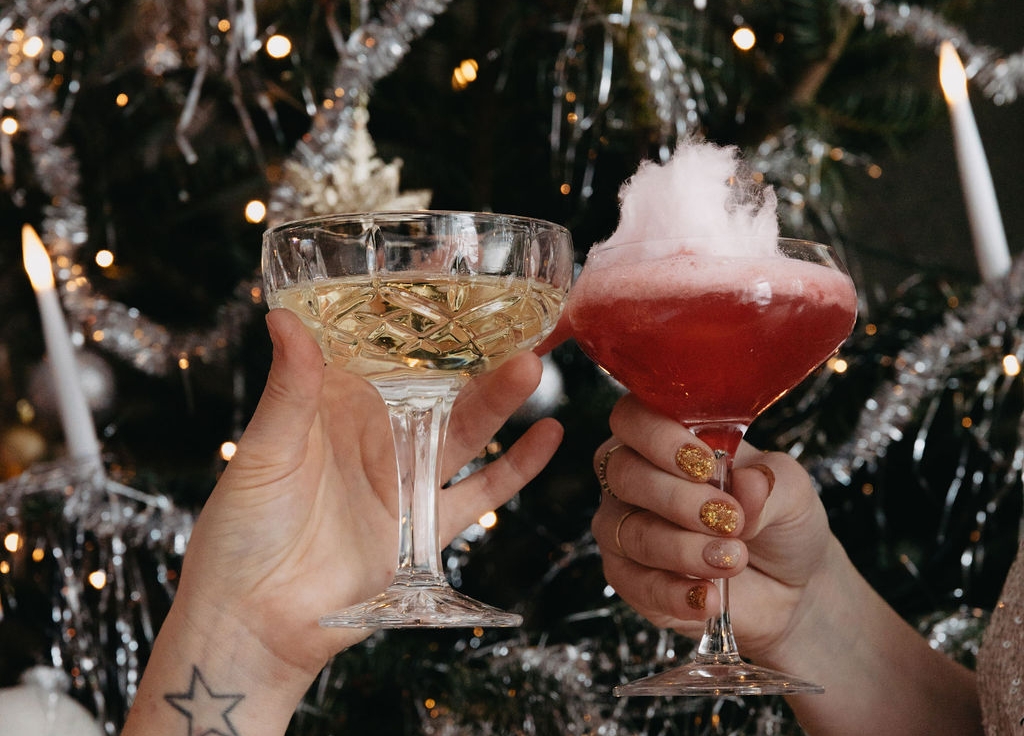 Två händer skålar med champagne och en röd sockervaddscocktail mot en julgran.