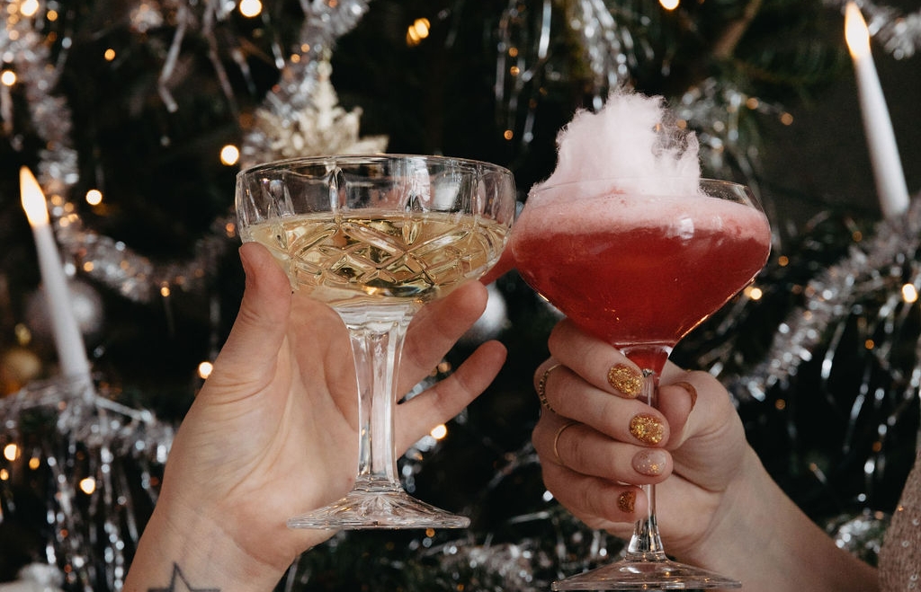 Två händer skålar med champagne och en röd sockervaddscocktail mot en julgran.