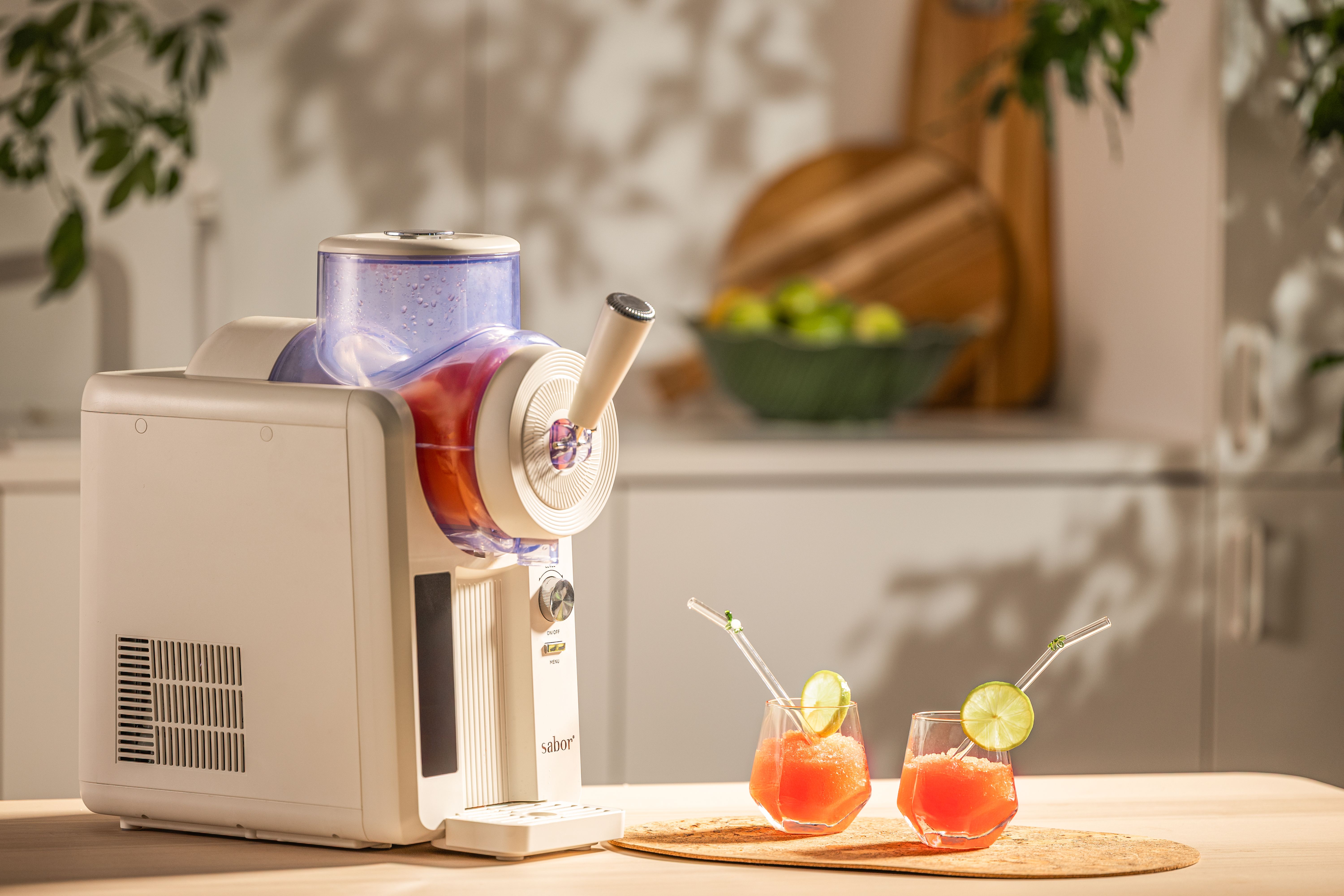 Valkoinen slushie-kone keittiön tiskillä kahden punaisen slushiedrinkin vieressä, koristeltuna limenviipaleilla ja pilleillä.