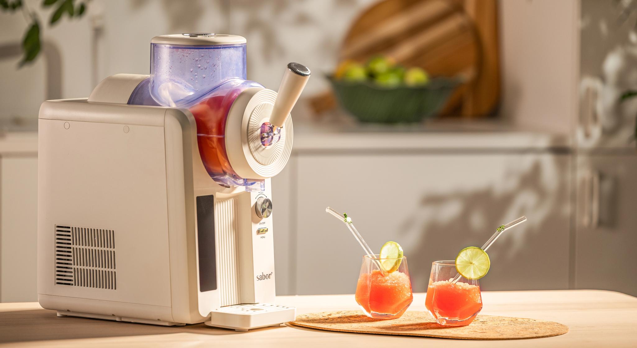 Valkoinen slushie-kone keittiön tiskillä kahden punaisen slushiedrinkin vieressä, koristeltuna limenviipaleilla ja pilleillä.